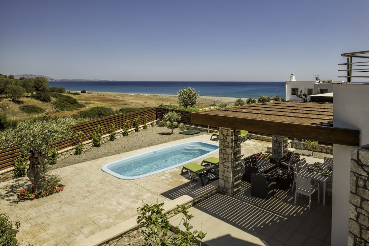 A rooftop pool area with a view of the sea, stone pillars, and outdoor furniture.
