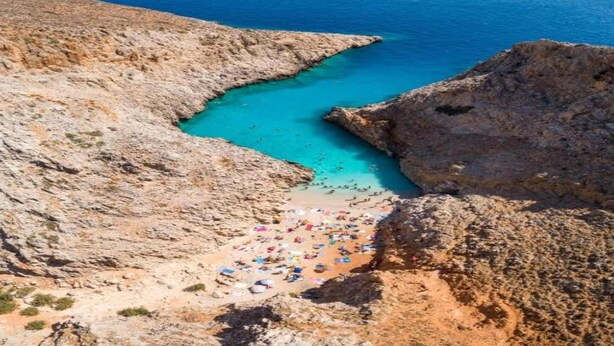 A rocky coastline with a clear blue sea, a narrow beach with colorful umbrellas, and a rocky cliff.