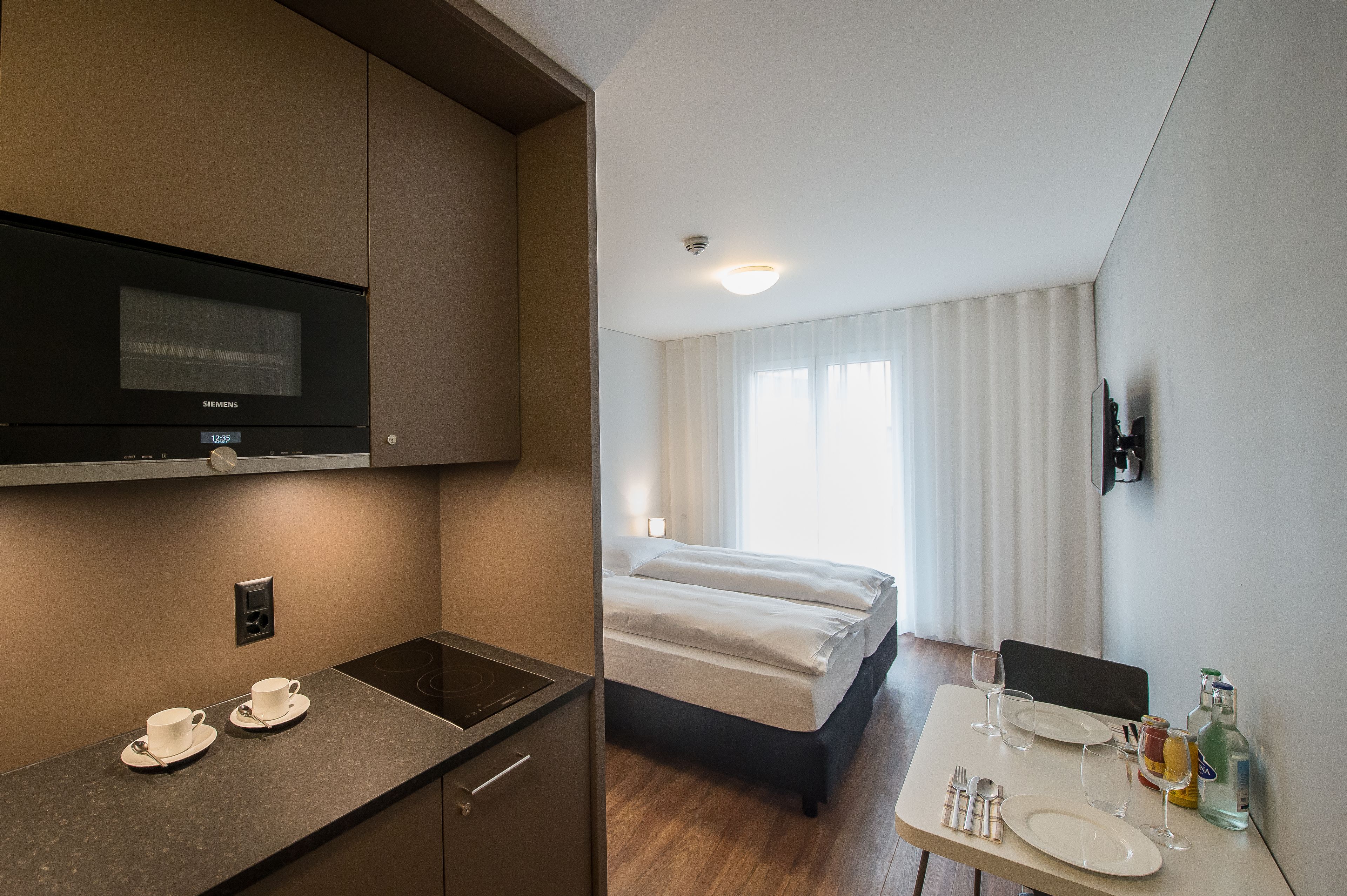 Une chambre d’hôtel équipée d’une kitchenette, d’un lit, d’un coin repas avec une table dressée pour deux personnes, et d’une grande fenêtre avec des rideaux.