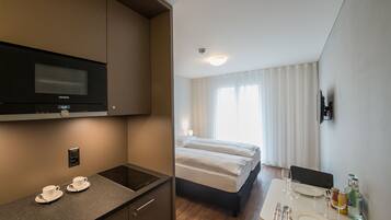 Une chambre d’hôtel équipée d’une kitchenette, d’un lit, d’un coin repas avec une table dressée pour deux personnes, et d’une grande fenêtre avec des rideaux.