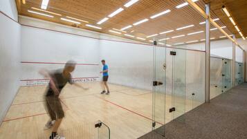 A squash court with wooden flooring and glass walls, a ceiling with linear lighting, and a visible number on the wall.