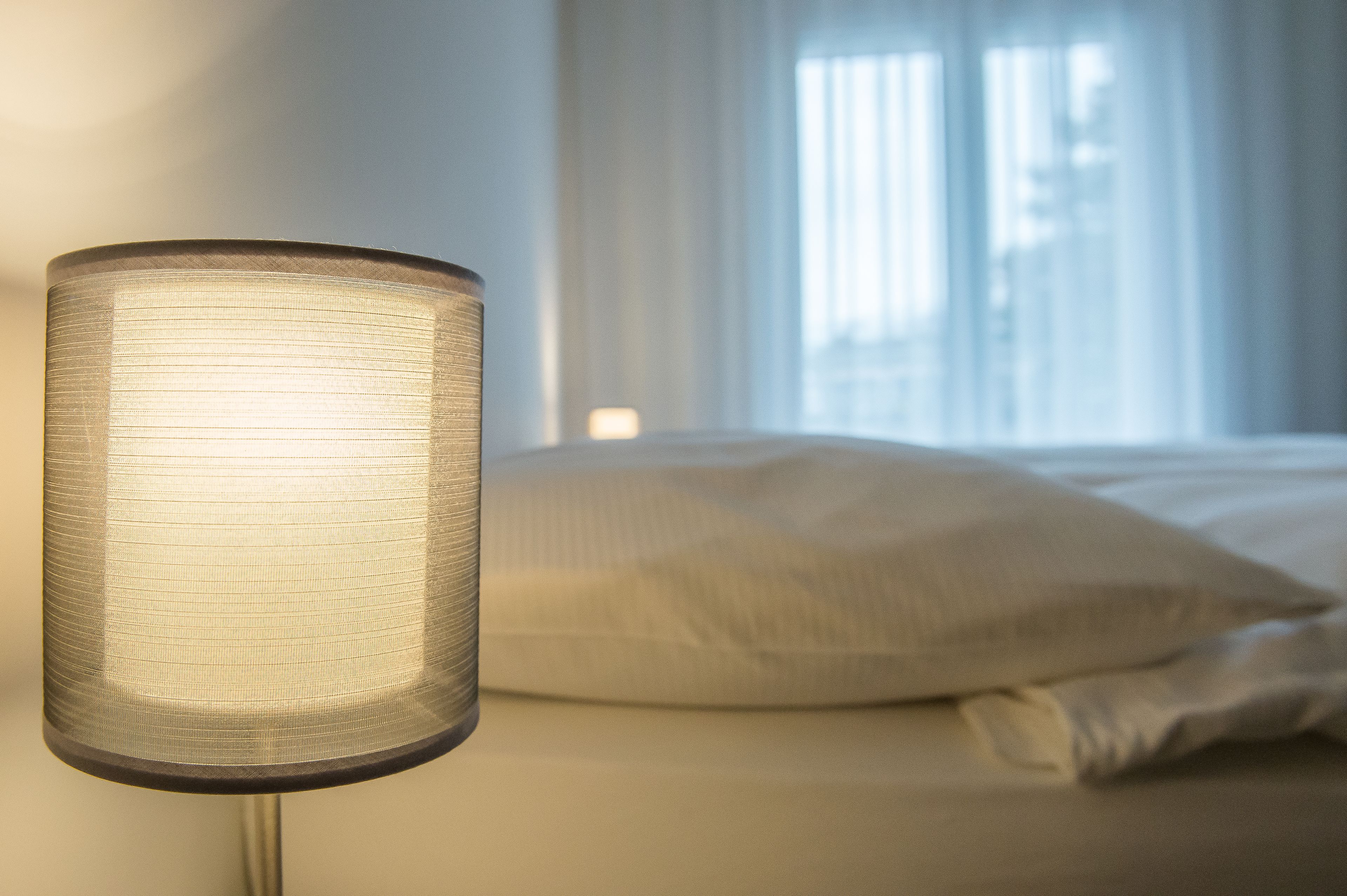A close-up of a cylindrical lamp with a textured shade, positioned in front of a bed with white bedding and a window with sheer curtains in the background.