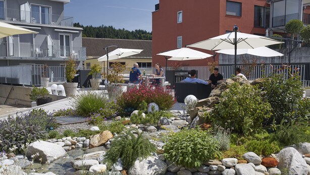 Un jardín con un estanque pequeño, rodeado de rocas y plantas. Hay mesas y sillas exteriores bajo sombrillas.