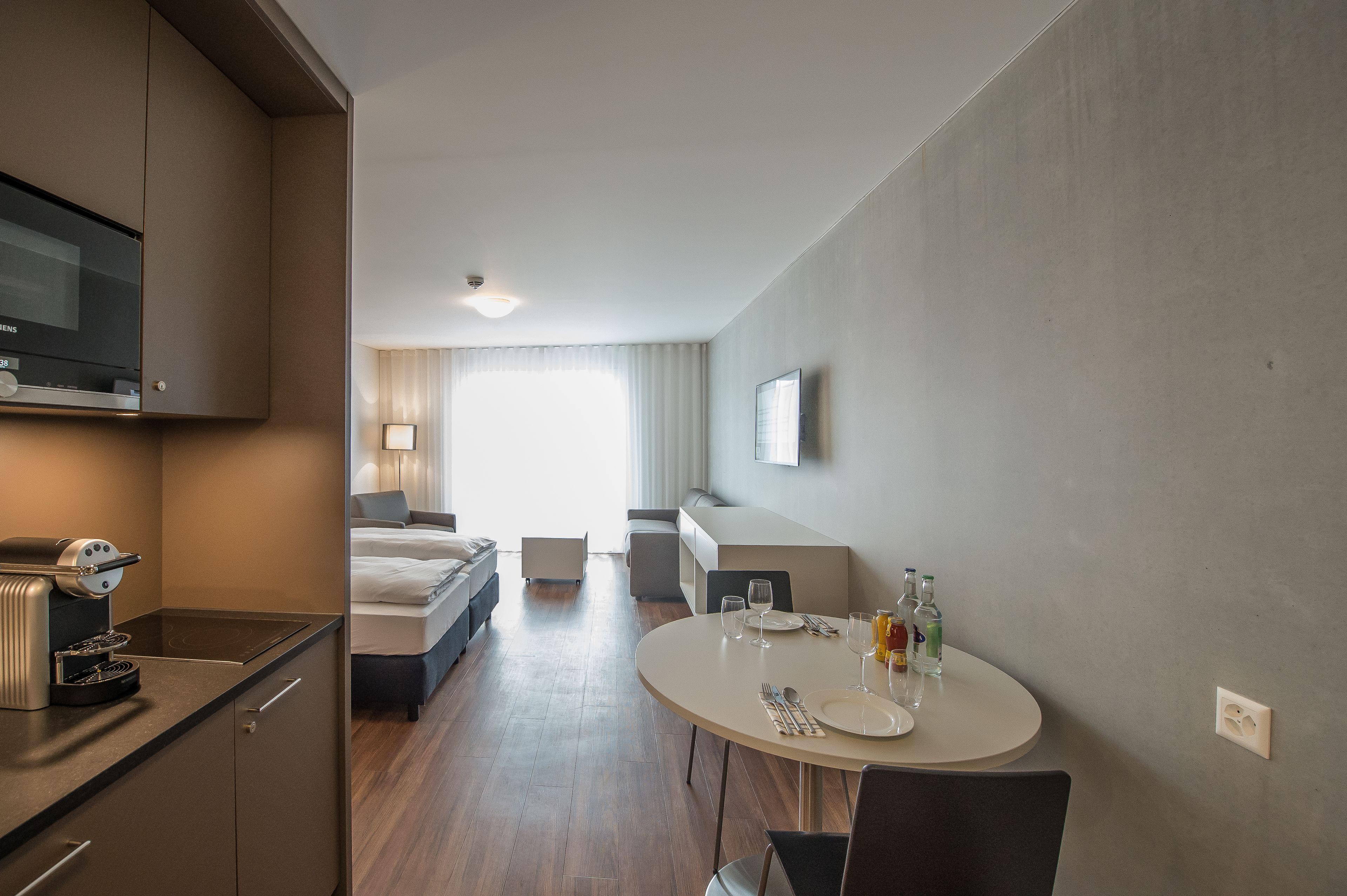 Una habitación de hotel con kitchenette, un comedor con mesa redonda, una cama, un sofá y un televisor montado en la pared.