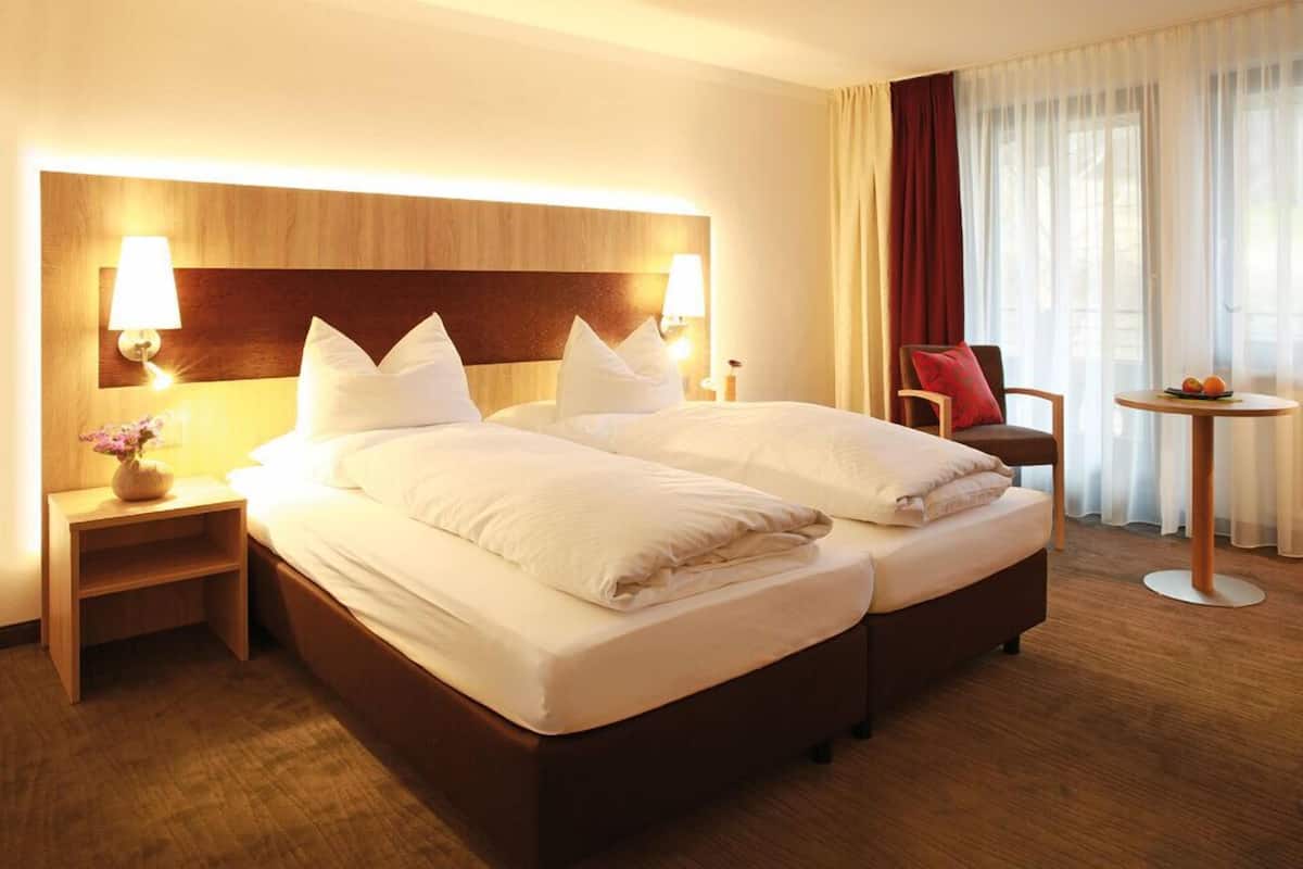 Ein Hotelzimmer mit einem großen Bett, zwei Nachttischen mit Lampen, einem Sessel, einem kleinen Tisch mit Obst und Blick ins Freie durch durchsichtigen Vorhängen.