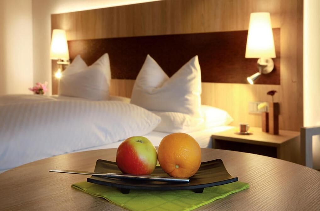 Ein Hotelzimmer mit einem ordentlich bezogenen Bett, zwei Nachttischlampen und einem Tisch mit einem Apfel und einer Orange.