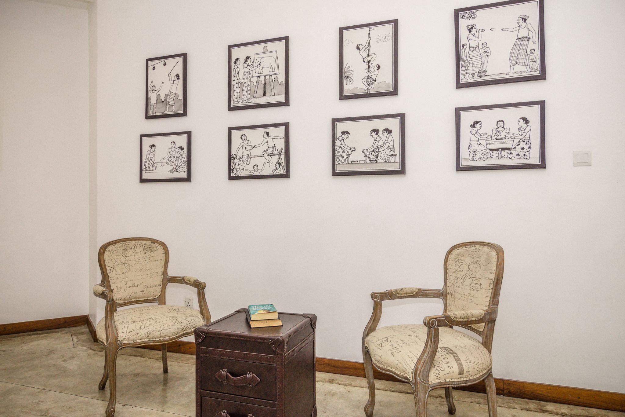 A gallery with framed black and white sketches on the wall and two antique chairs with a small table between them.