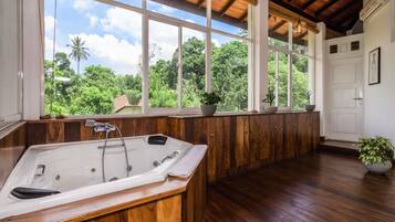 A spacious bathroom with a large bathtub, wooden flooring, and a view of lush greenery through large windows.