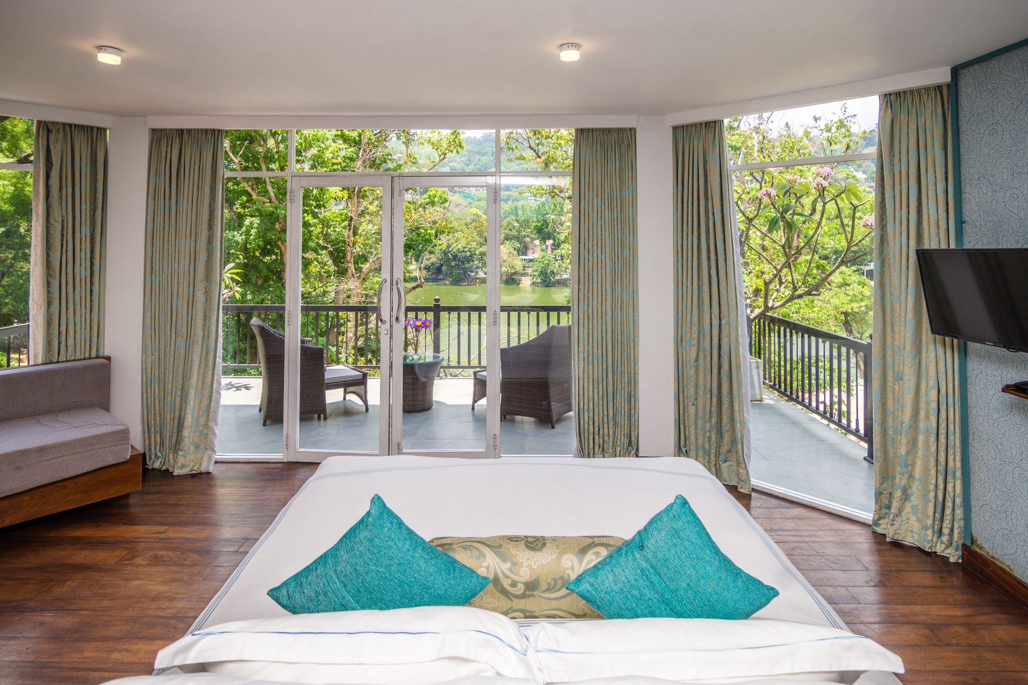 A bedroom with a large bed, a balcony with wicker furniture, and a TV mounted on the wall.