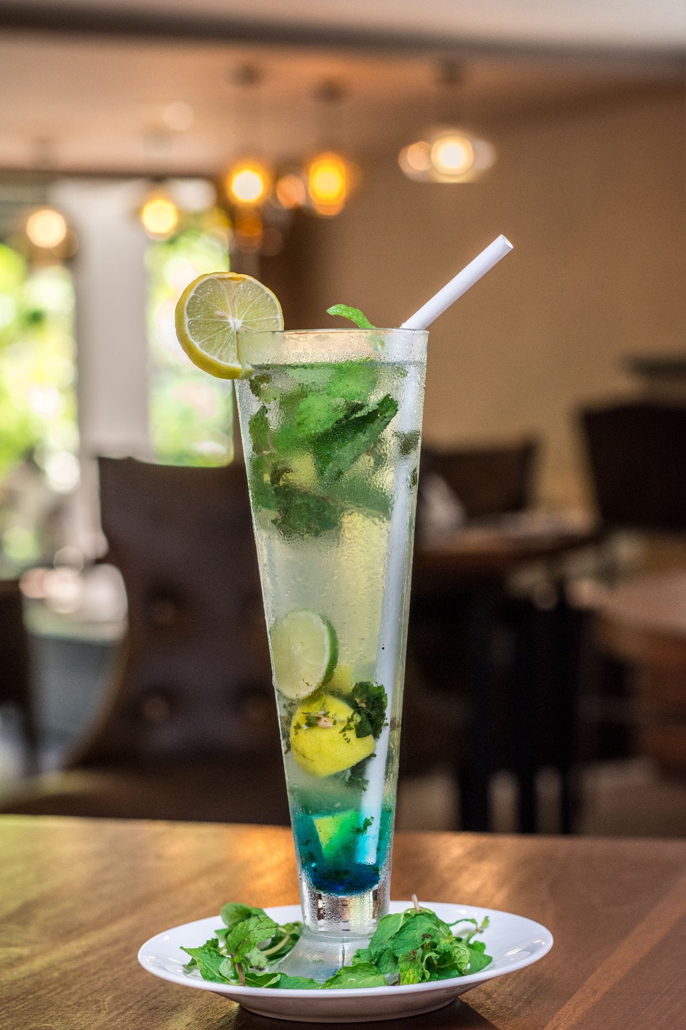 Een hoog glas met een blauwe drank, gesneden limoen en muntblaadjes, geplaatst op een wit bord met extra munt op een houten tafel.