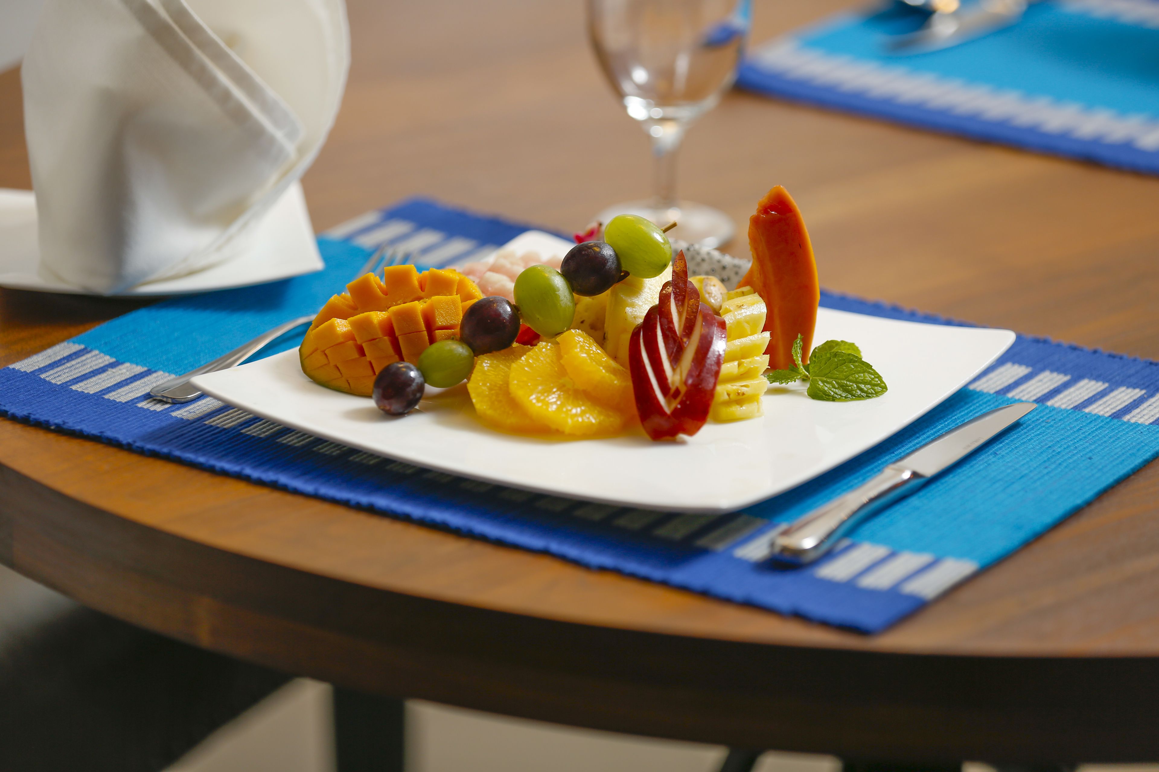 A fruit platter on a white plate with a blue placemat, a glass of water, and a white pitcher on a wooden table.