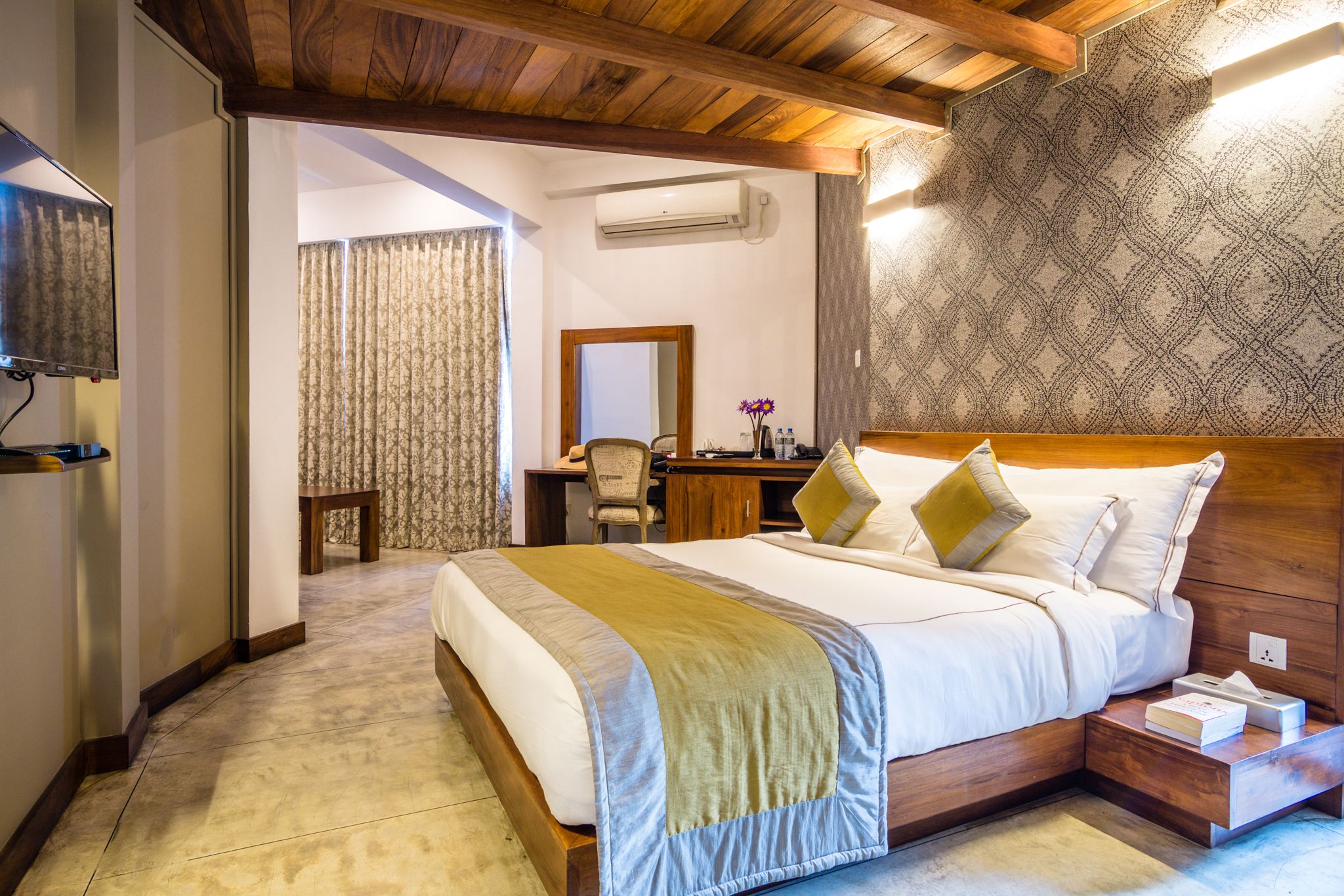 Quarto de hotel com teto de madeira, uma cama grande com cobertor bege e verde, uma escrivaninha com cadeira e parede estampada.
