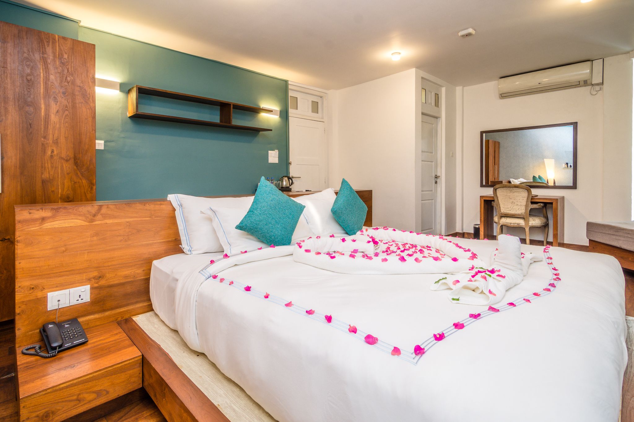 A hotel room with a wooden headboard, a desk with a chair, a mirror, and a bed with a white bedspread and teal pillows.