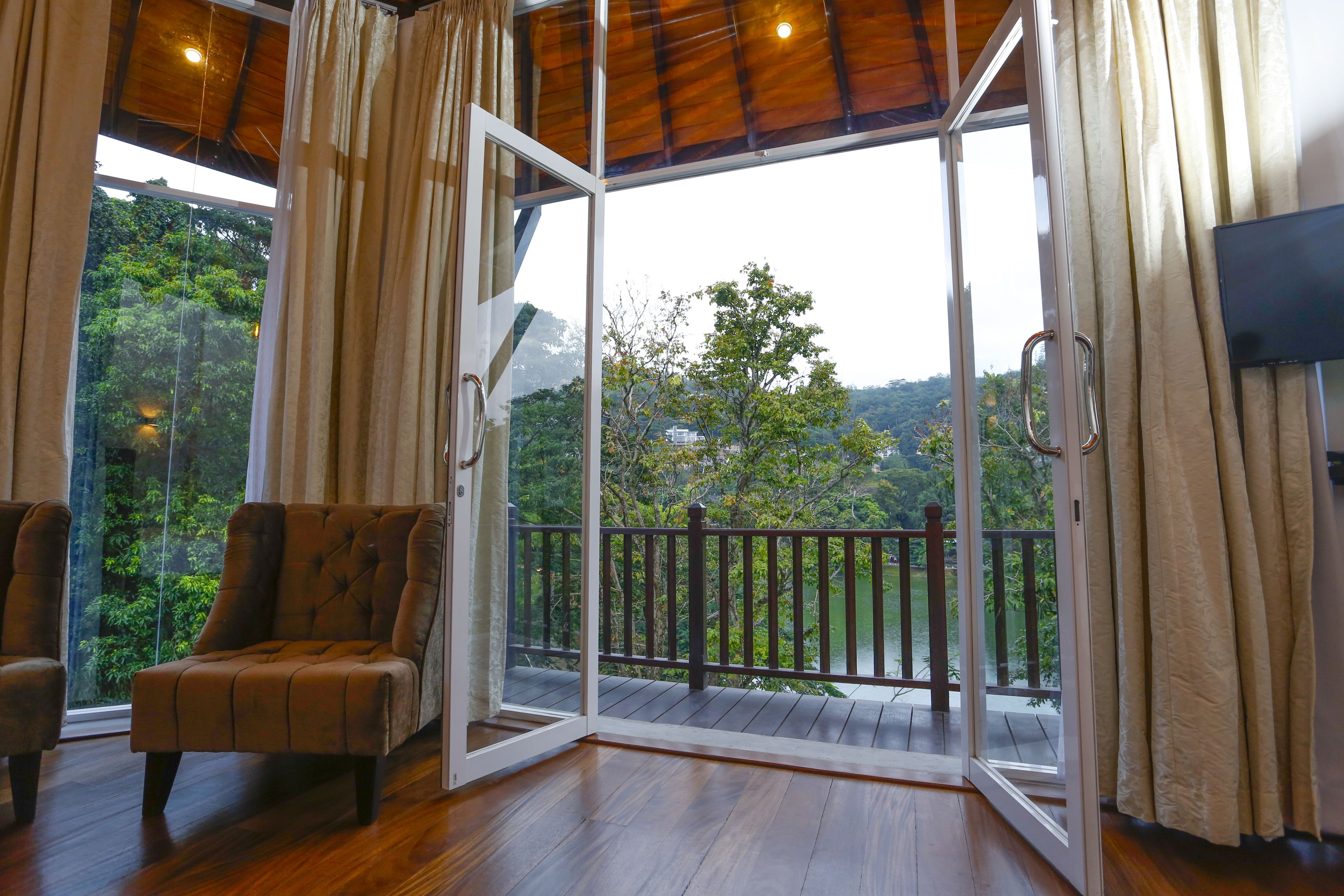 Een kamer met een houten vloer, een bruine fauteuil, een schuifdeur van glas naar een balkon met houten balustrade, en een aan de muur bevestigde televisie.