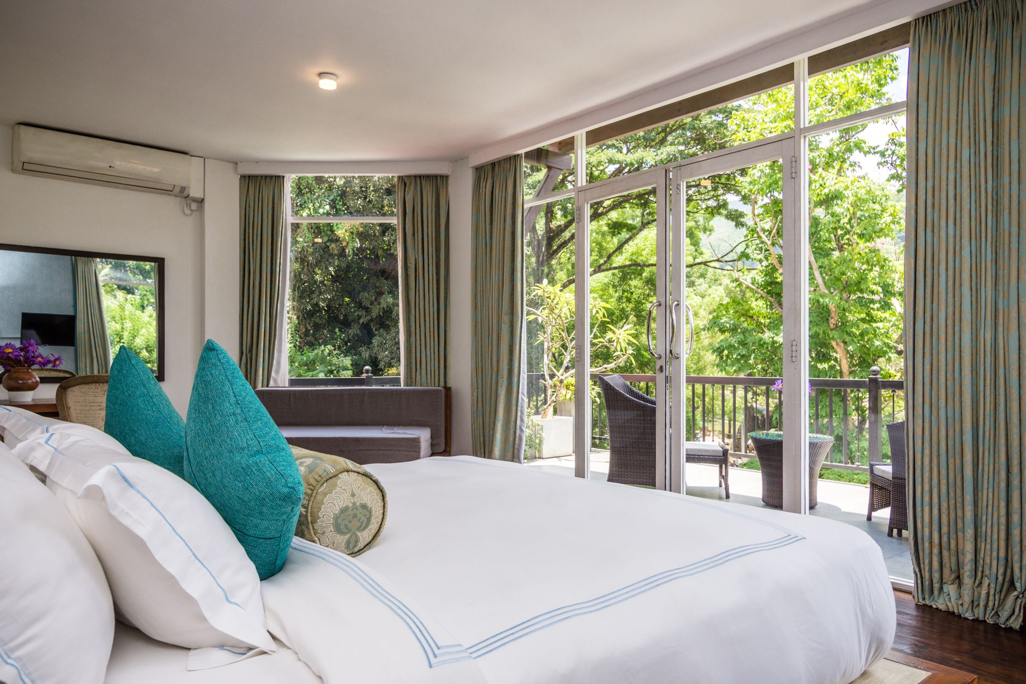 A spacious bedroom with a large bed, a television, and a balcony with wicker furniture.