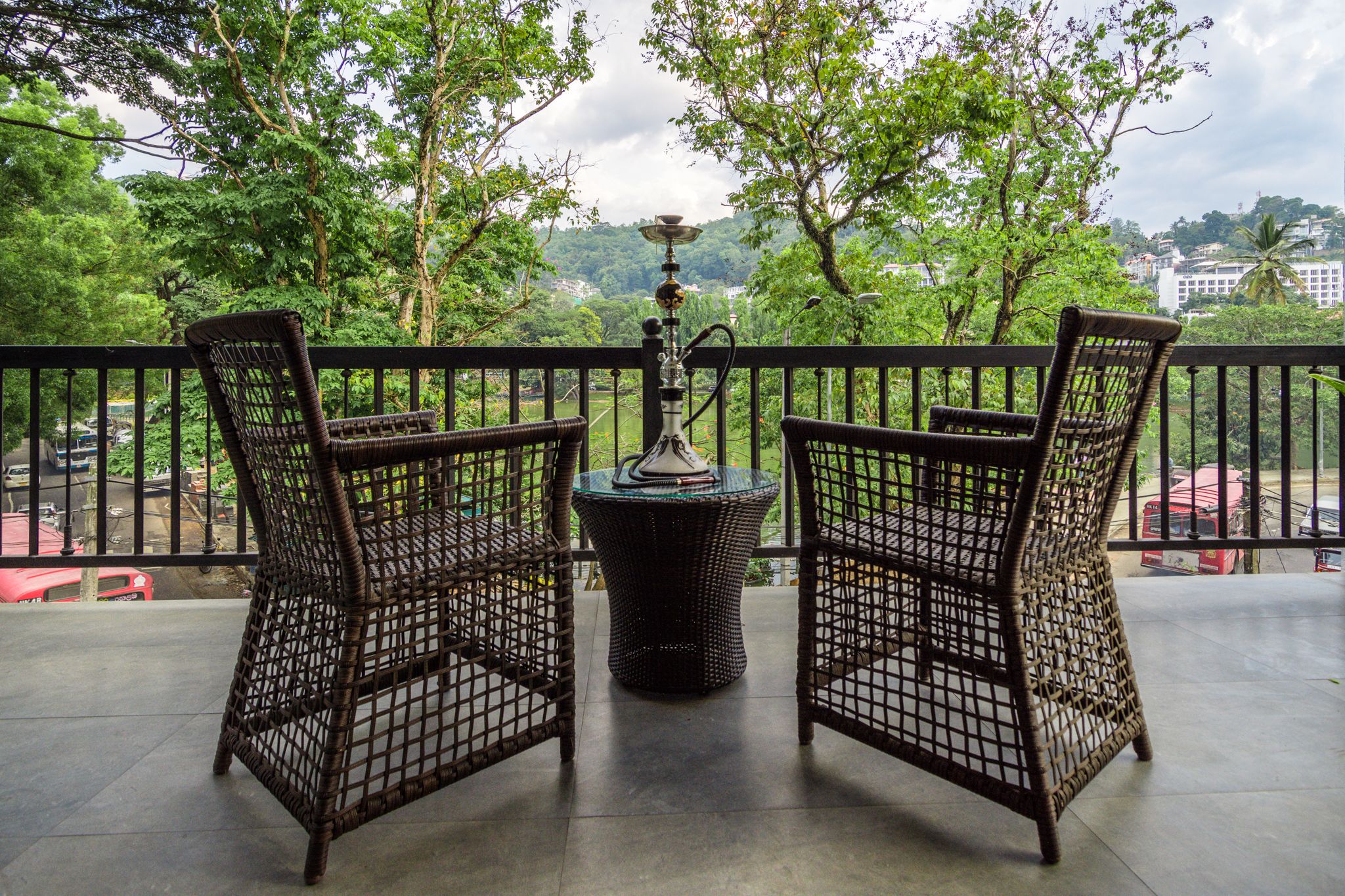 A balcony with wicker chairs and a table, overlooking a lush green landscape and a distant cityscape.
