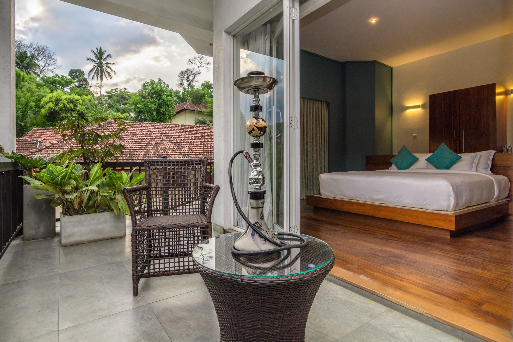 A balcony with a wicker chair, a glass table, and a hookah. The room has a bed with white bedding and teal pillows.