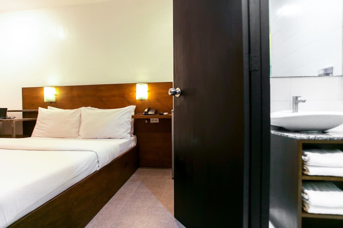 Une chambre d’hôtel avec une tête de lit en bois, un lit avec du linge de maison blanc, une table de chevet avec une lampe et une salle de bain équipée d’un lavabo et d’un porte-serviettes.