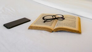 A neatly made bed with a book and glasses on it, a bedside table, and a wall-mounted light. - Hotel Durban (Makati)