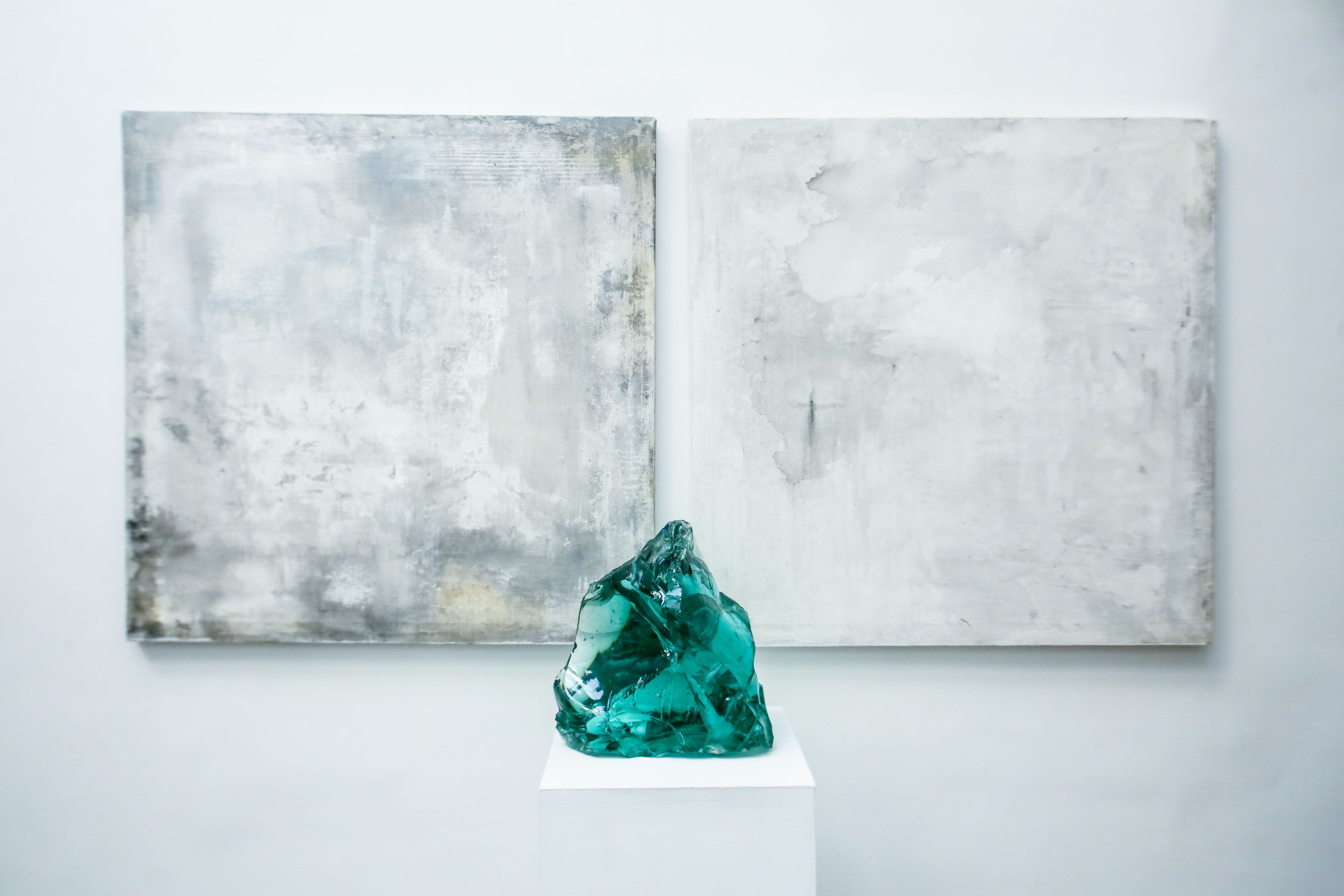 A teal crystal sculpture on a pedestal against a white wall with two abstract paintings.