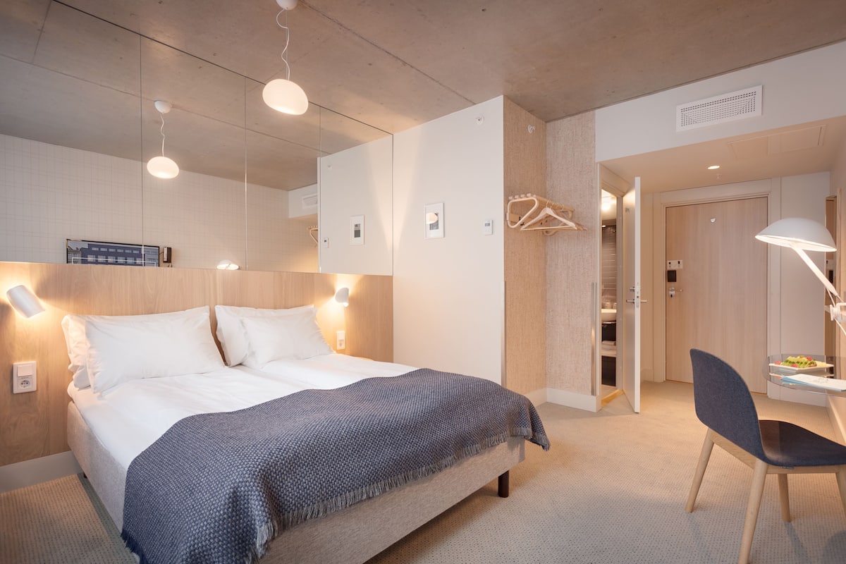 A modern hotel room with a large bed, bedside lamps, a desk with a chair, and a wardrobe.