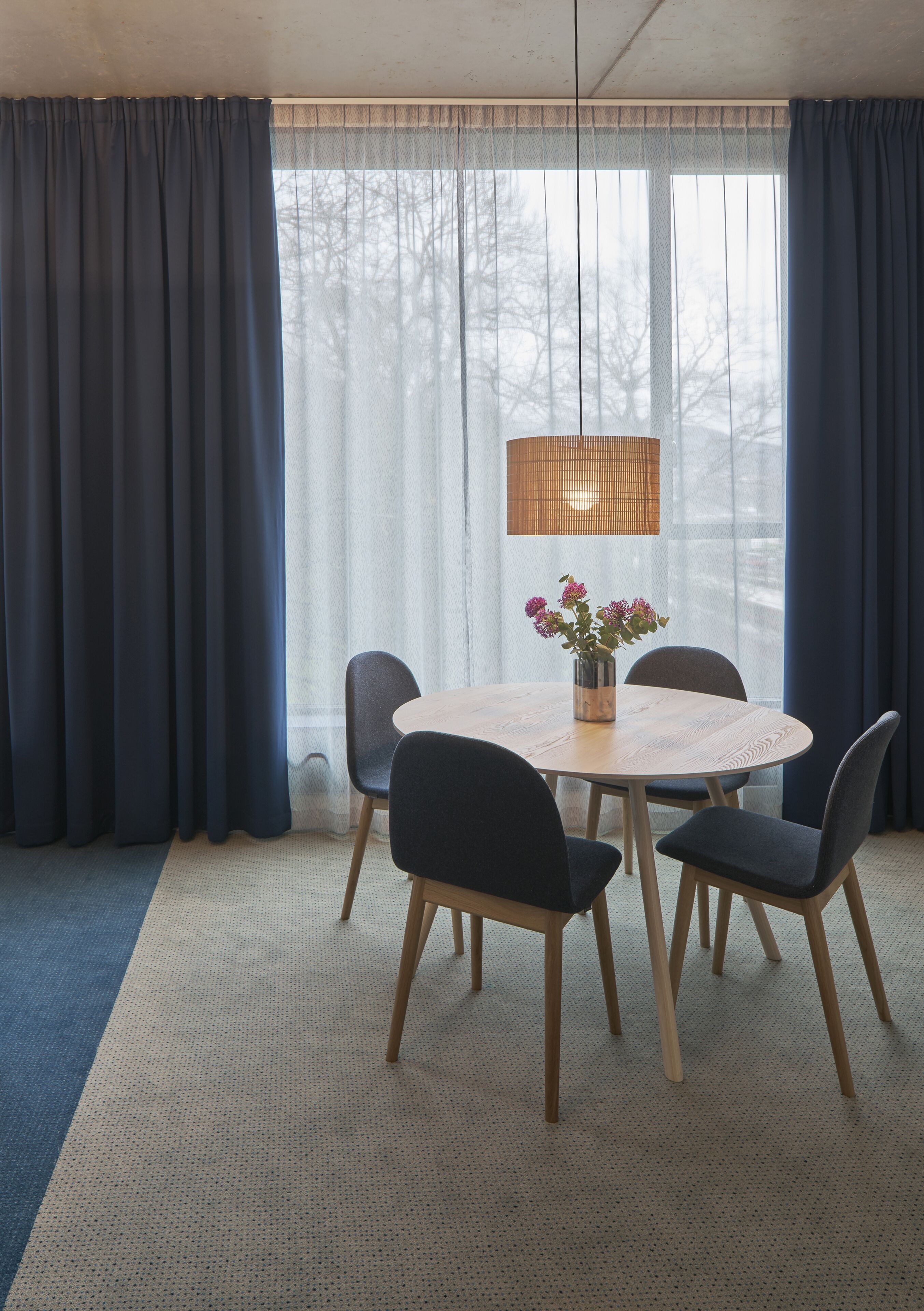 A dining area with a round table, four chairs, a vase with flowers, and a hanging lamp.