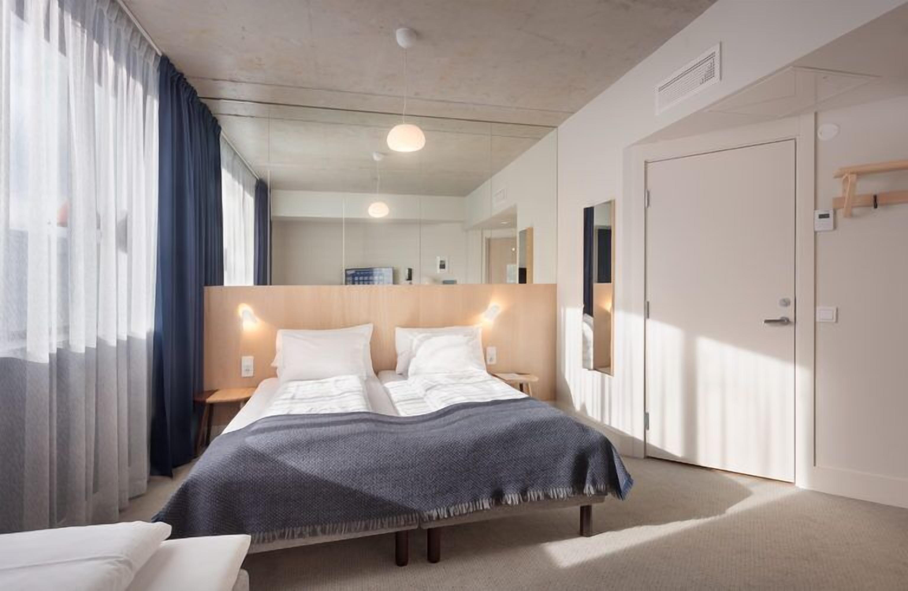 A modern hotel room with a large bed, a bedside table, a nightstand, a wardrobe, and a mirror.