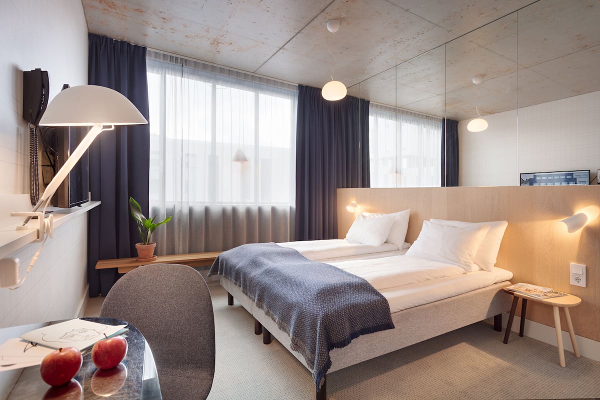 A hotel room with a bed, bedside table, lamp, and a desk with a chair and a plant.