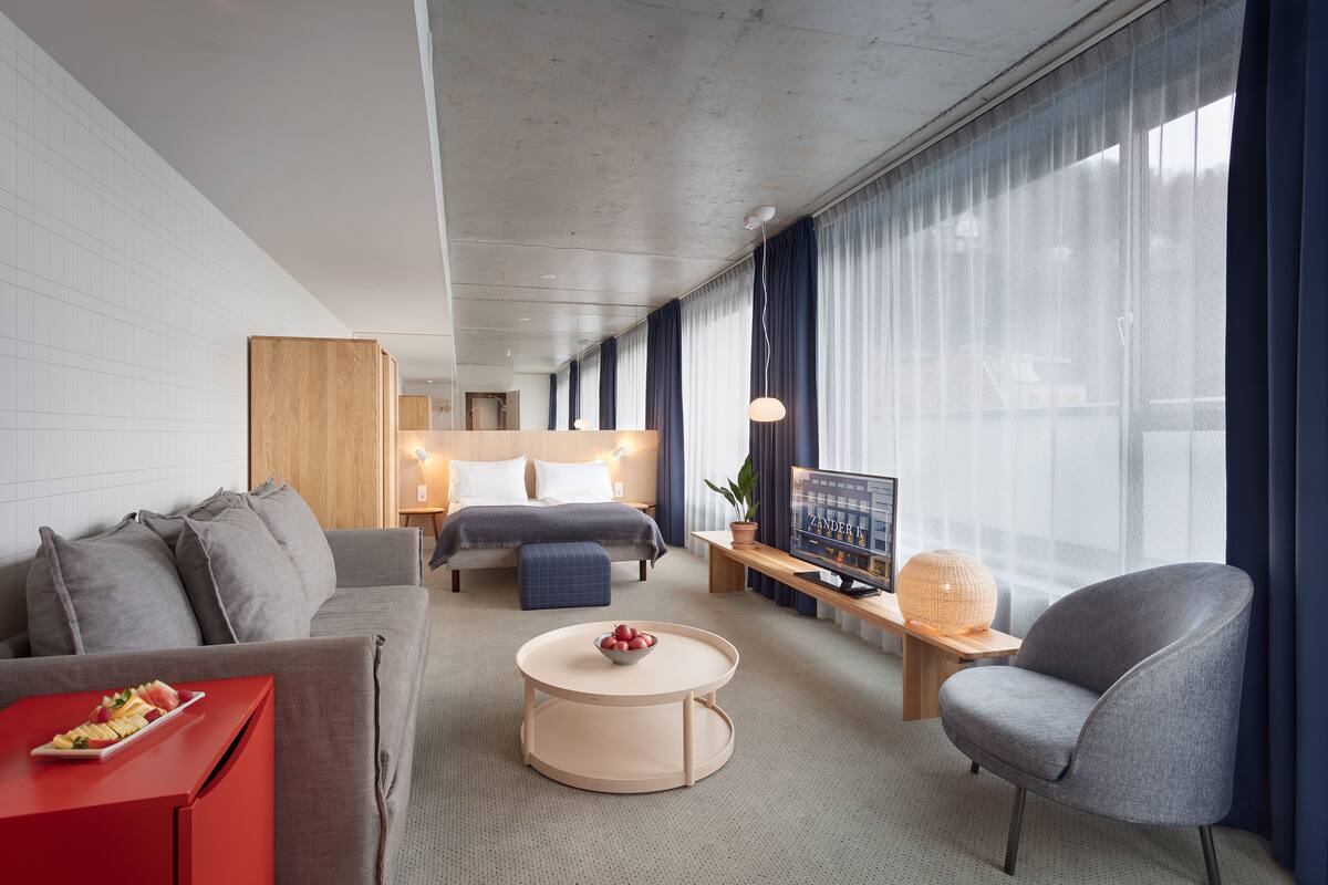 A modern hotel room with a grey sofa, a red side table, a round coffee table, a grey armchair, a bed with a wooden headboard, a bedside table with a lamp, a large window with curtains, and a television on a wooden stand.