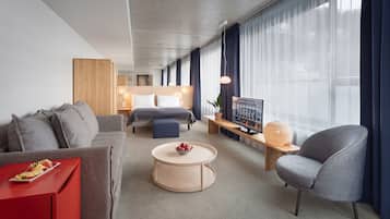 Een moderne hotelkamer met een grijze bank, een rode bijzettafel, een ronde salontafel, een grijze fauteuil, een bed met een houten hoofdbord, een nachtkastje met een lamp, een groot raam met gordijnen en een televisie op een houten dressoir.