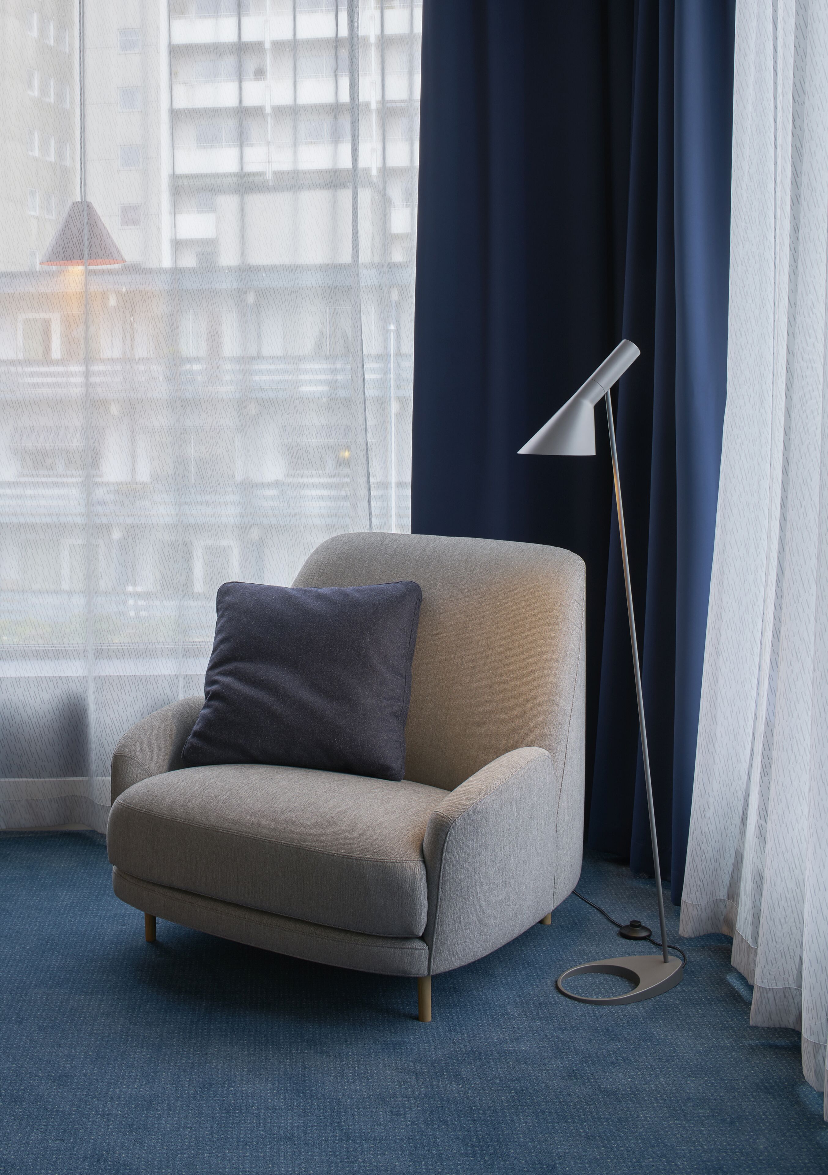 A modern armchair with a cushion, positioned near a window with sheer curtains, a floor lamp, and a blue carpet.