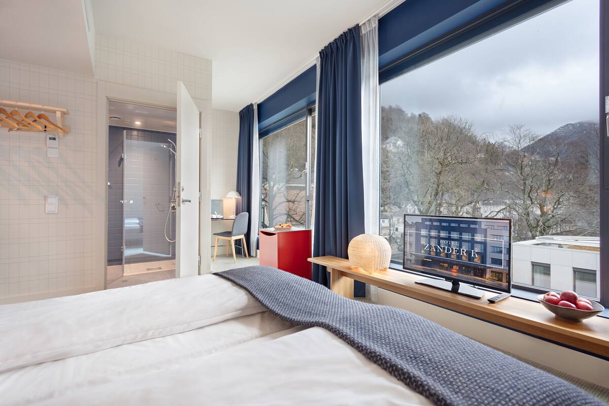 A hotel room with a bed, a desk with a computer, a window with a view of trees and mountains, and a shower area.