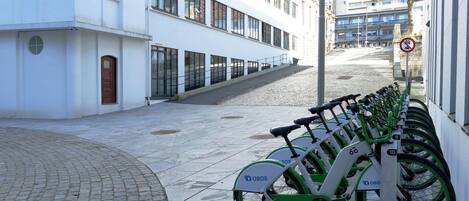 Una fila de bicicletas OBOS, verdes y blancas, estacionadas a lo largo de una acera junto a un edificio blanco con ventanas amplias.