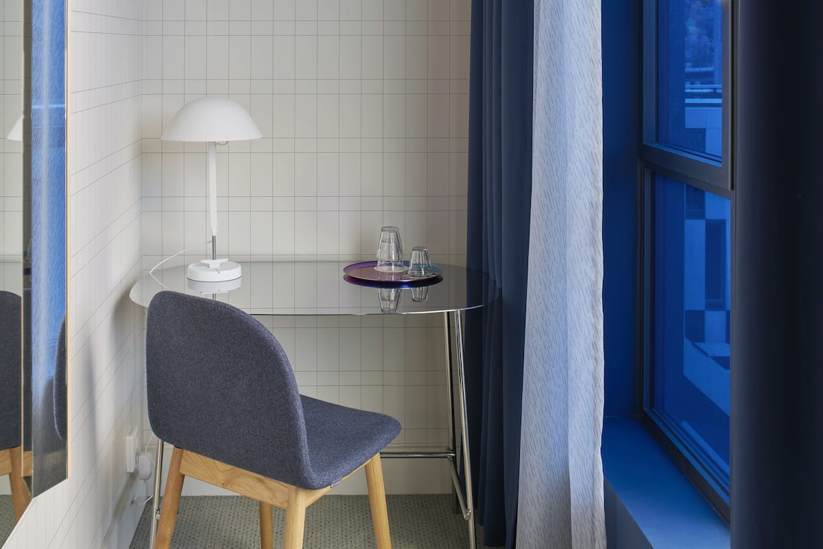 A modern bathroom with a chair, a white lamp, a small round table with a purple top, and a window with blue curtains.