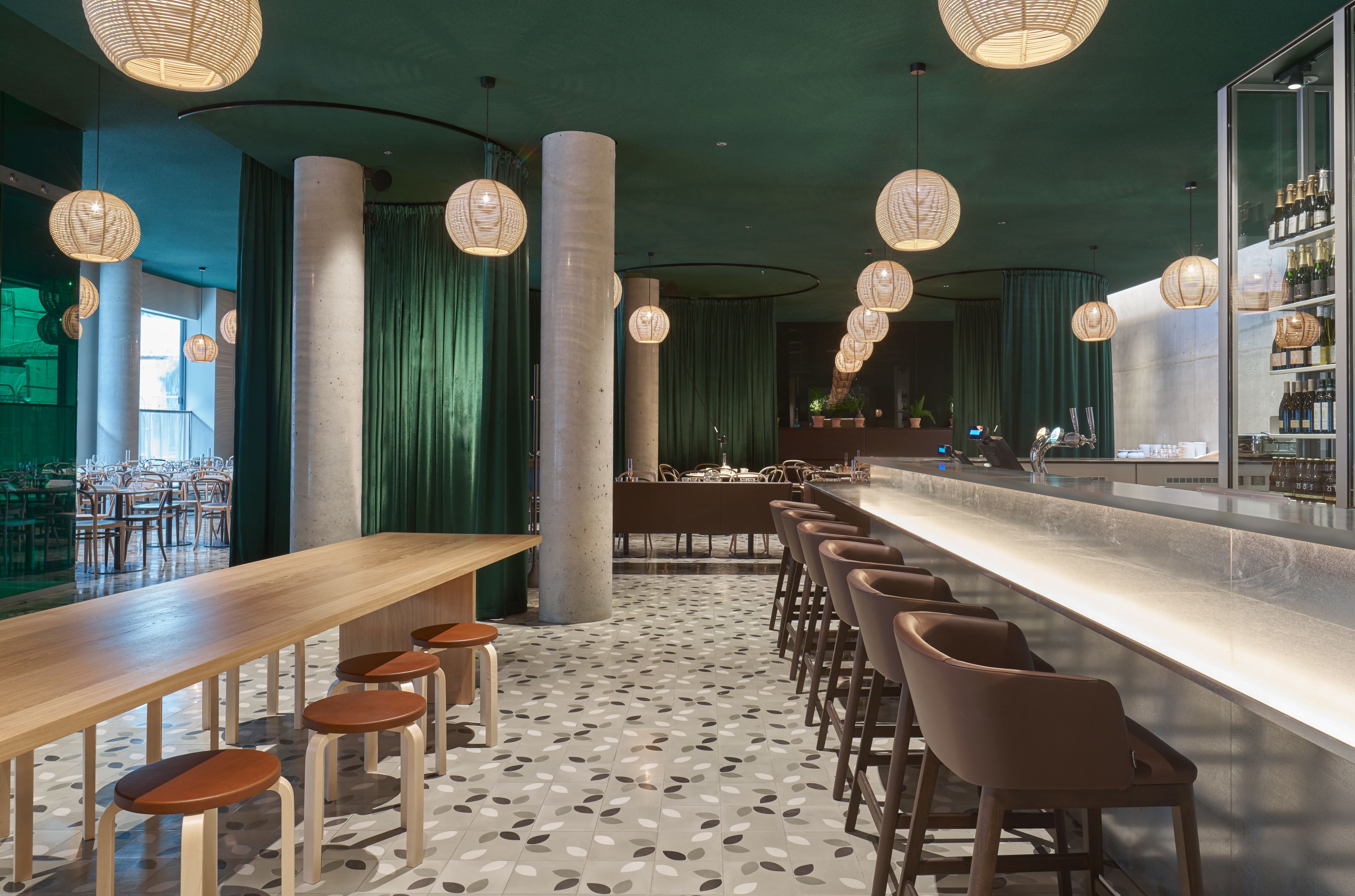 Zona de bar moderna con mesas y taburetes de madera, una barra alargada y lámparas decorativas colgantes.