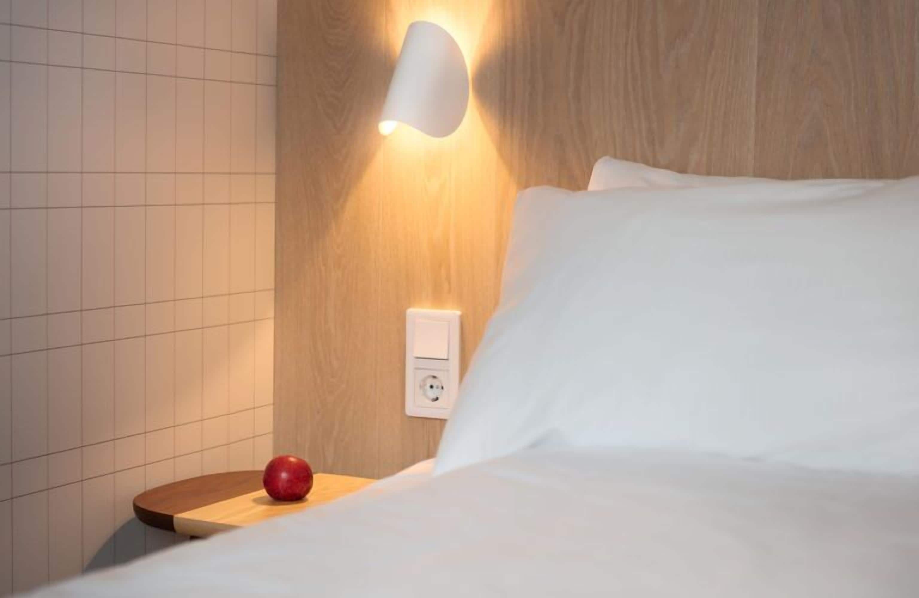 Una cama bien hecha con sábanas blancas, una mesita de noche de madera con una manzana roja, una lámpara de pared y un enchufe.