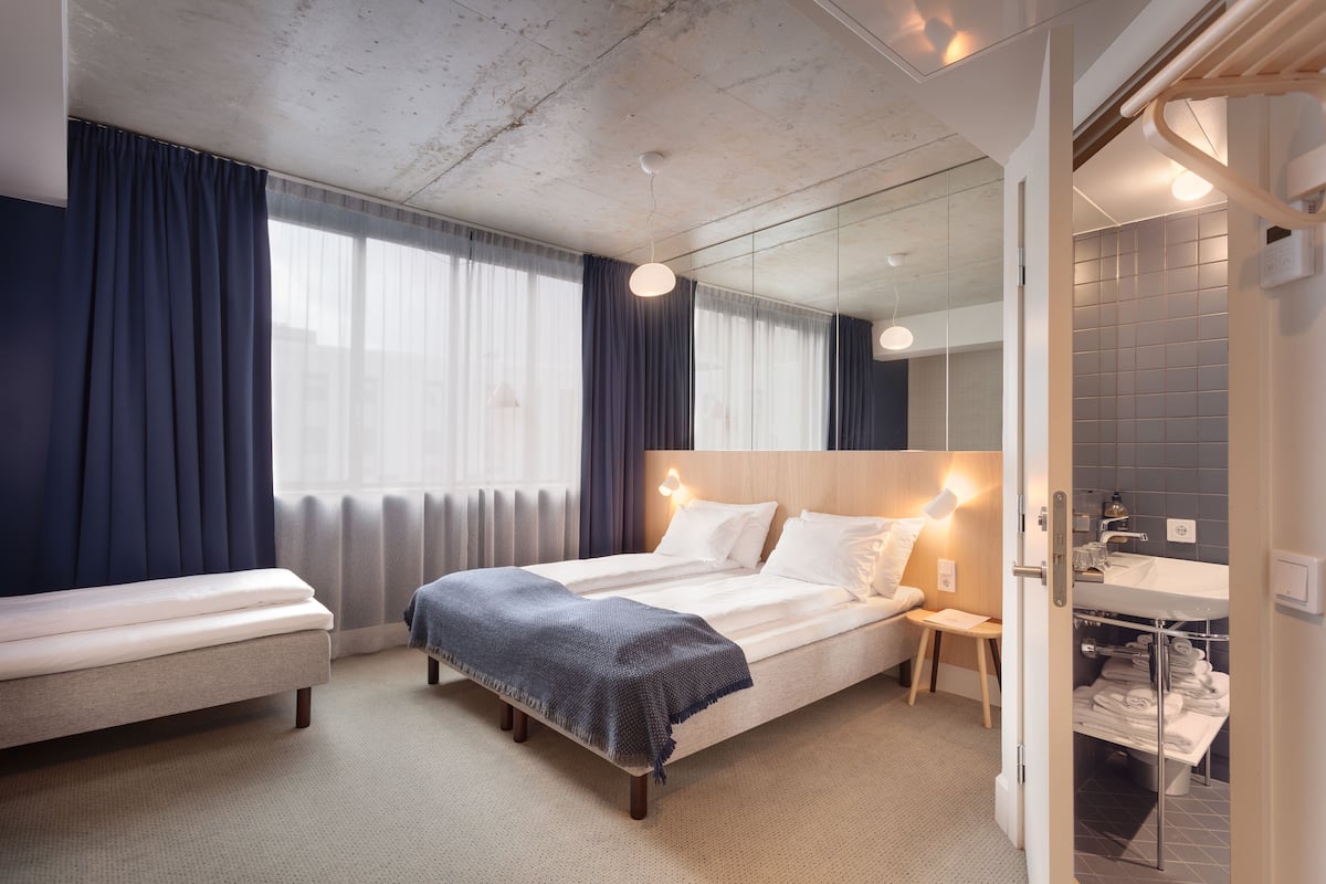 A modern hotel room with a large bed, bedside tables, a mirror, and a bathroom with a sink and towel rack.