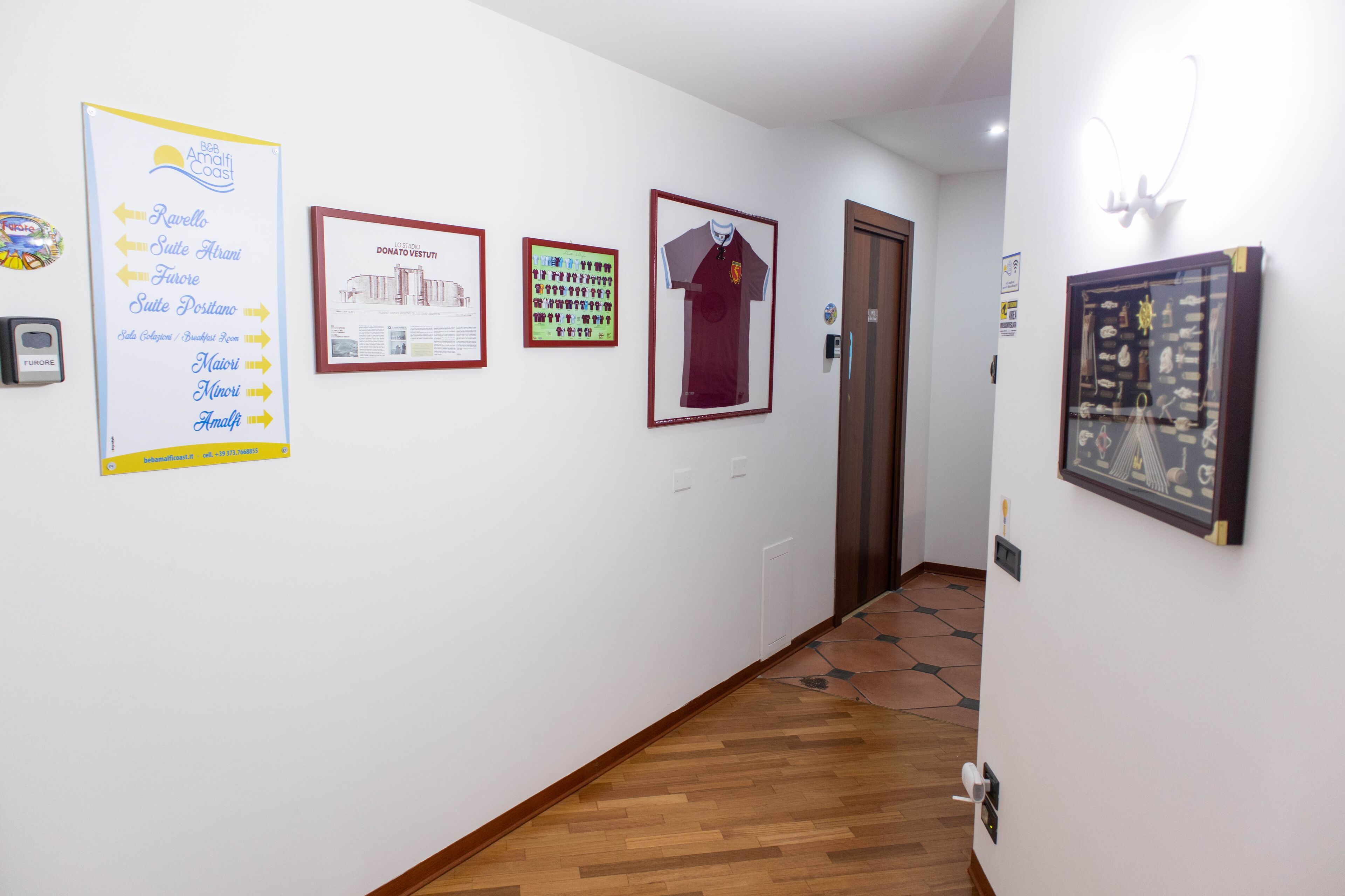 Un corridoio con pavimento in legno, pareti bianche e diverse opere d'arte appese, tra cui una mappa, una maglia sportiva e un quadro incorniciato.