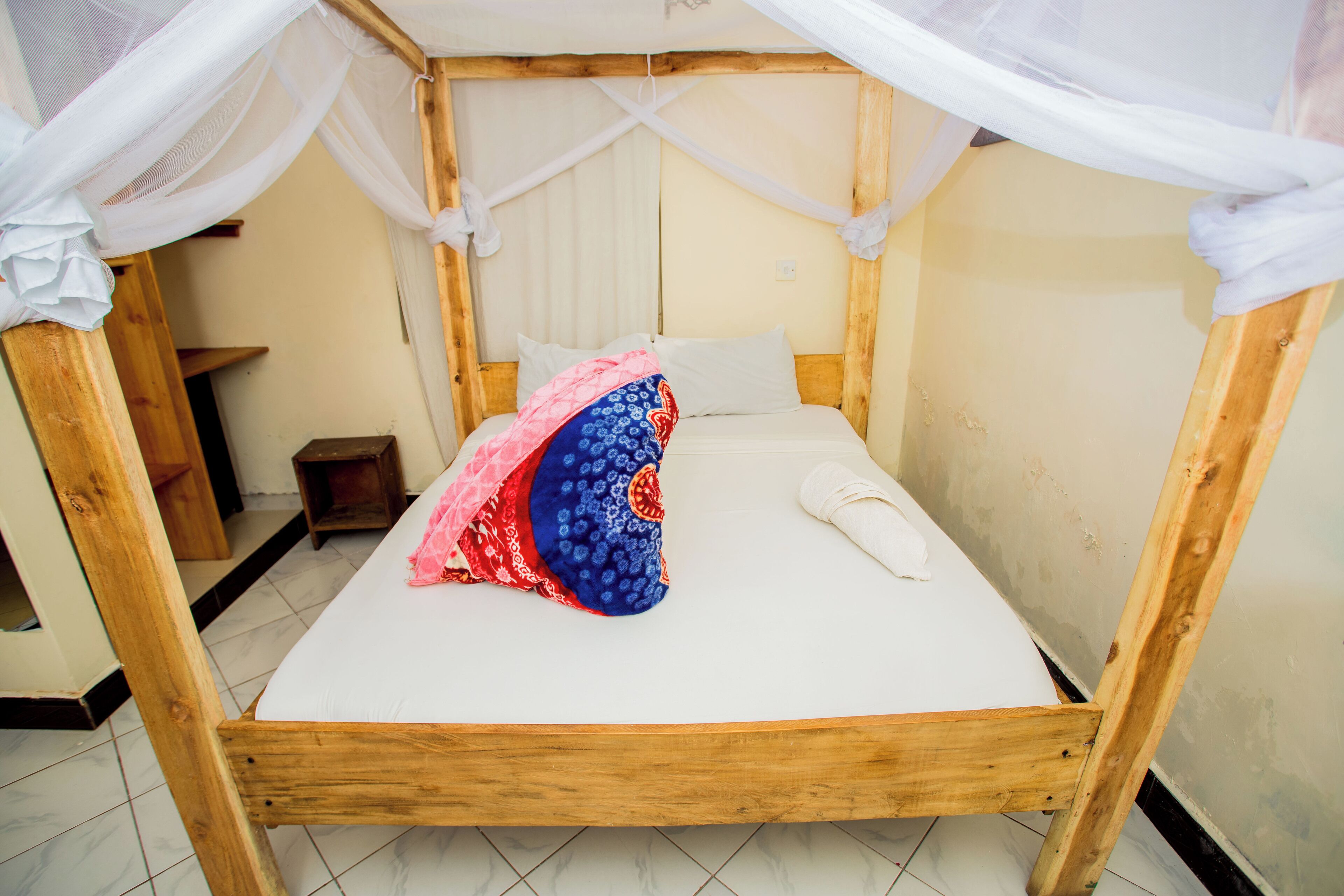 Een houten bed met een witte matras, een blauw-rood kussen en een witte handdoek. Er staat een houten nachtkastje naast het bed.