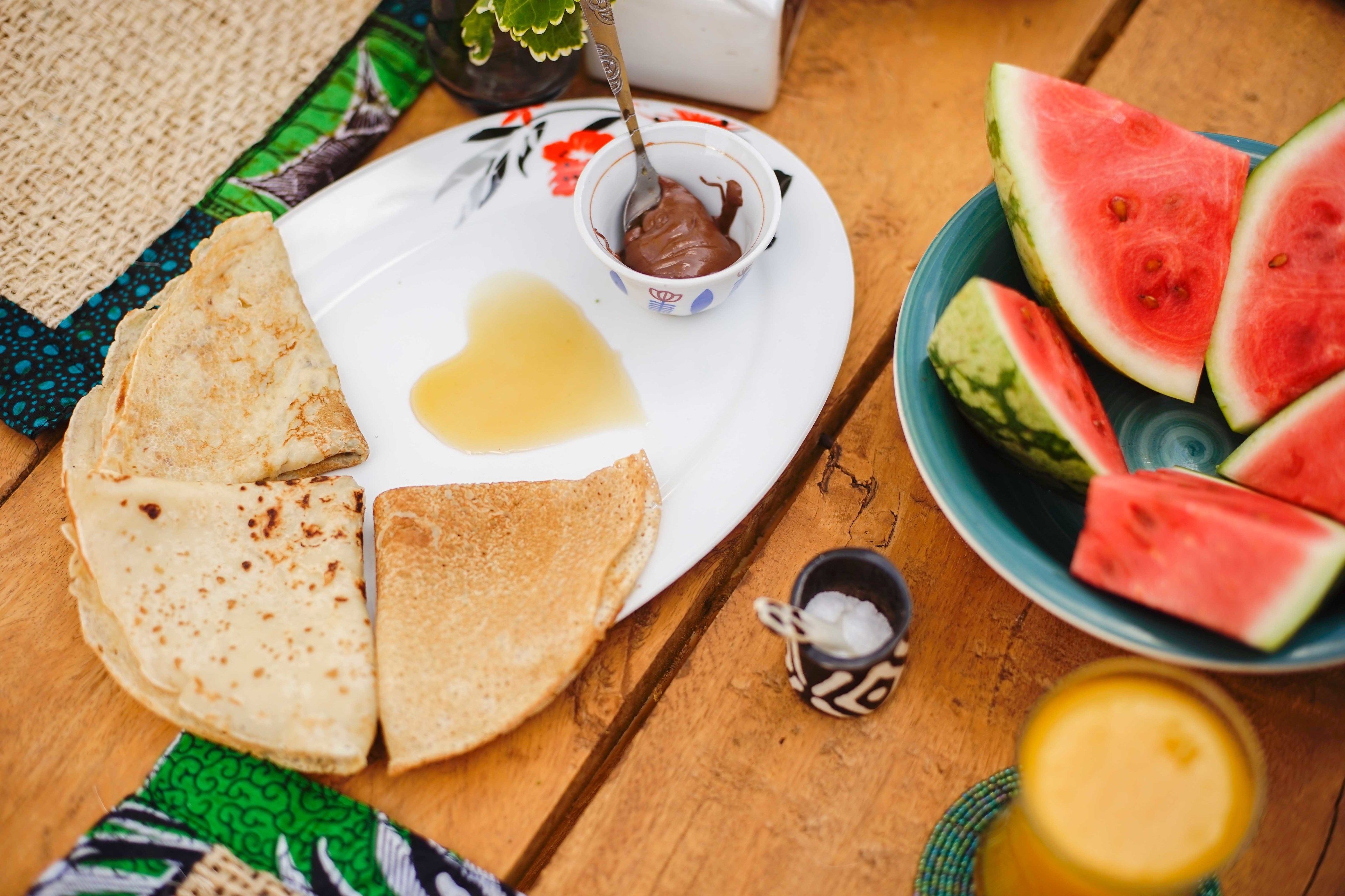 A plate with crepes, a small bowl of chocolate spread, and slices of watermelon on a wooden table.