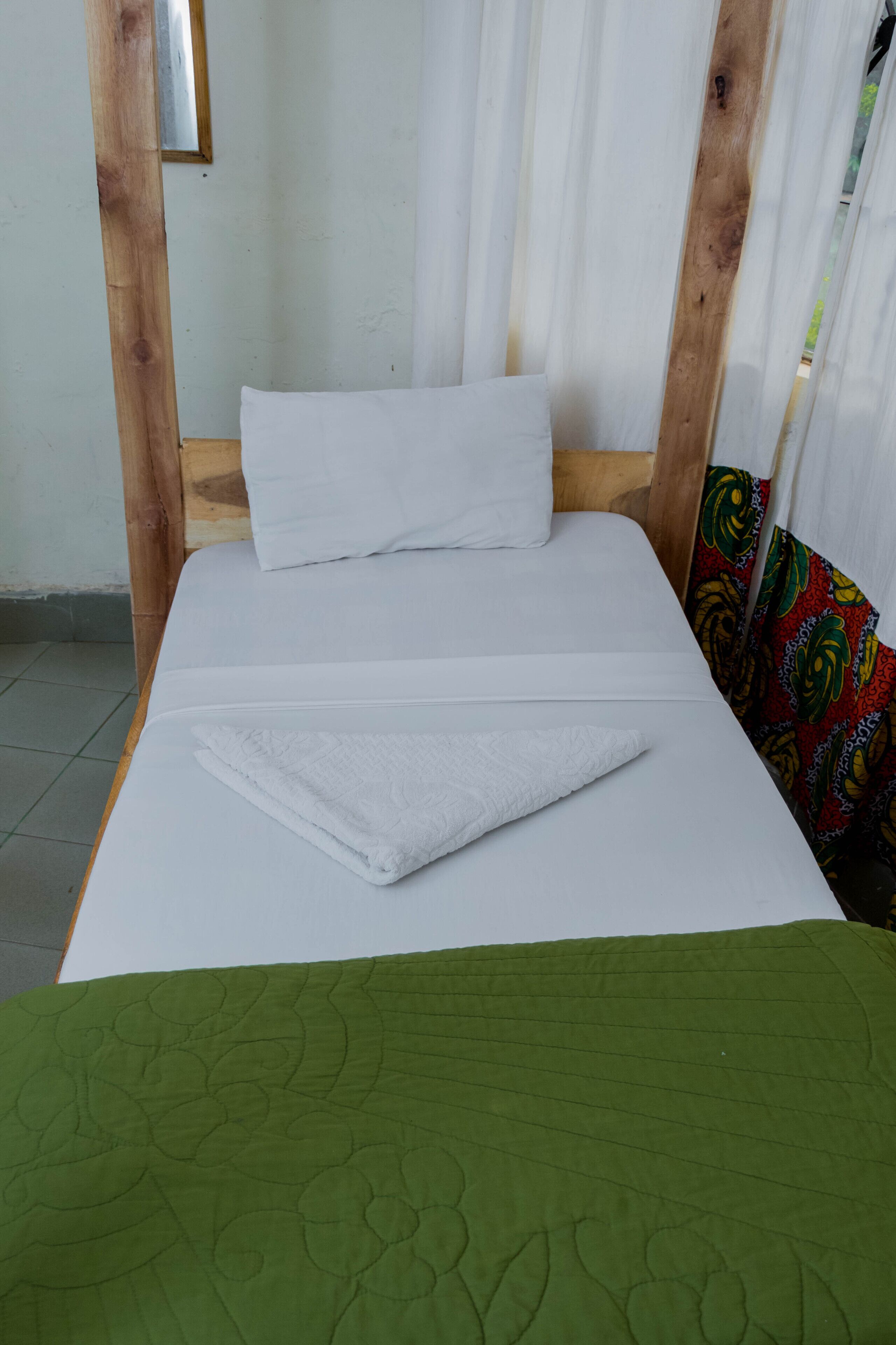A single bed with white bedding and a green quilt, wooden headboard, and a colorful curtain.