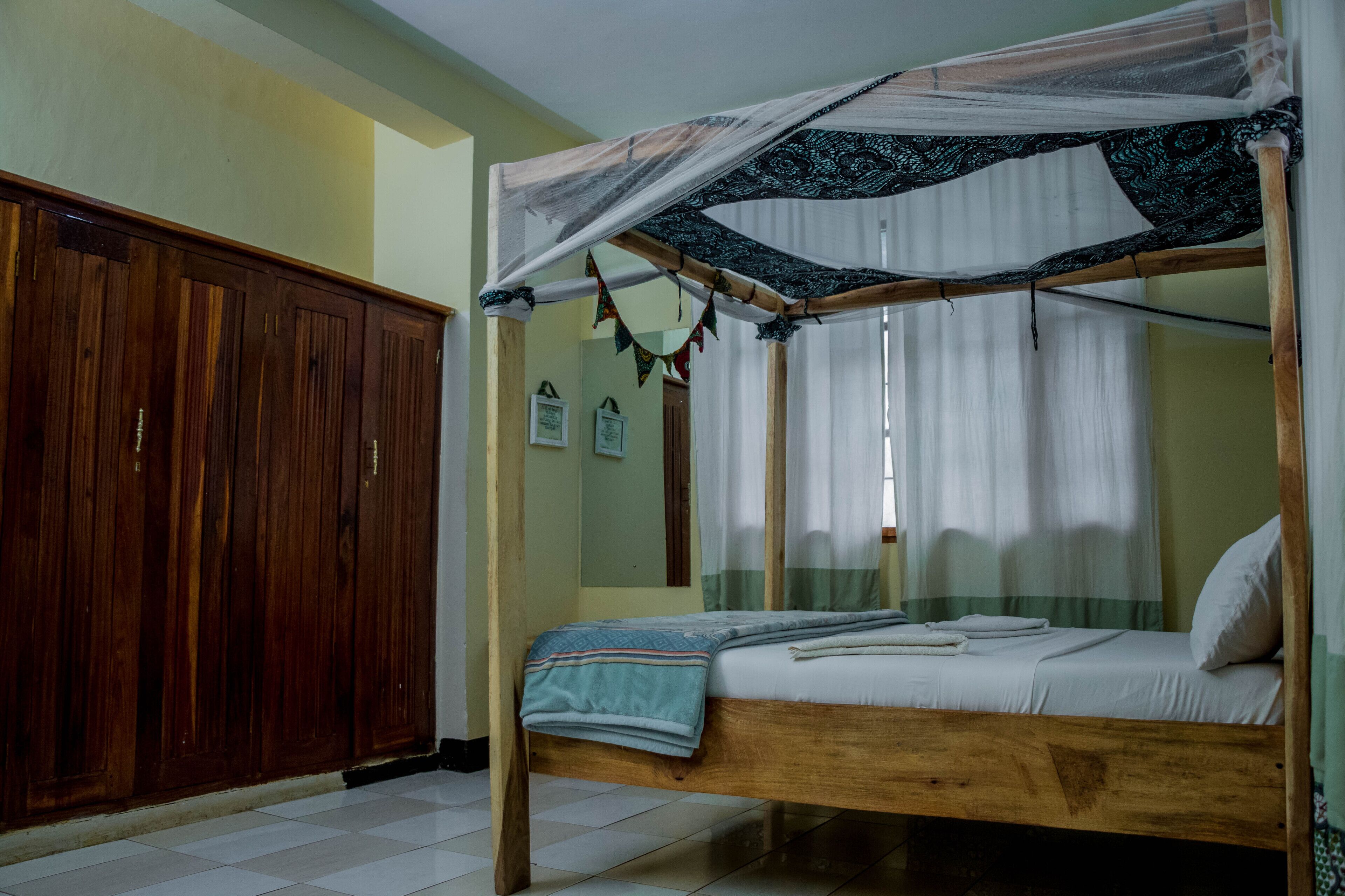 A single bed with a mosquito net, wooden wardrobe, and tiled floor.