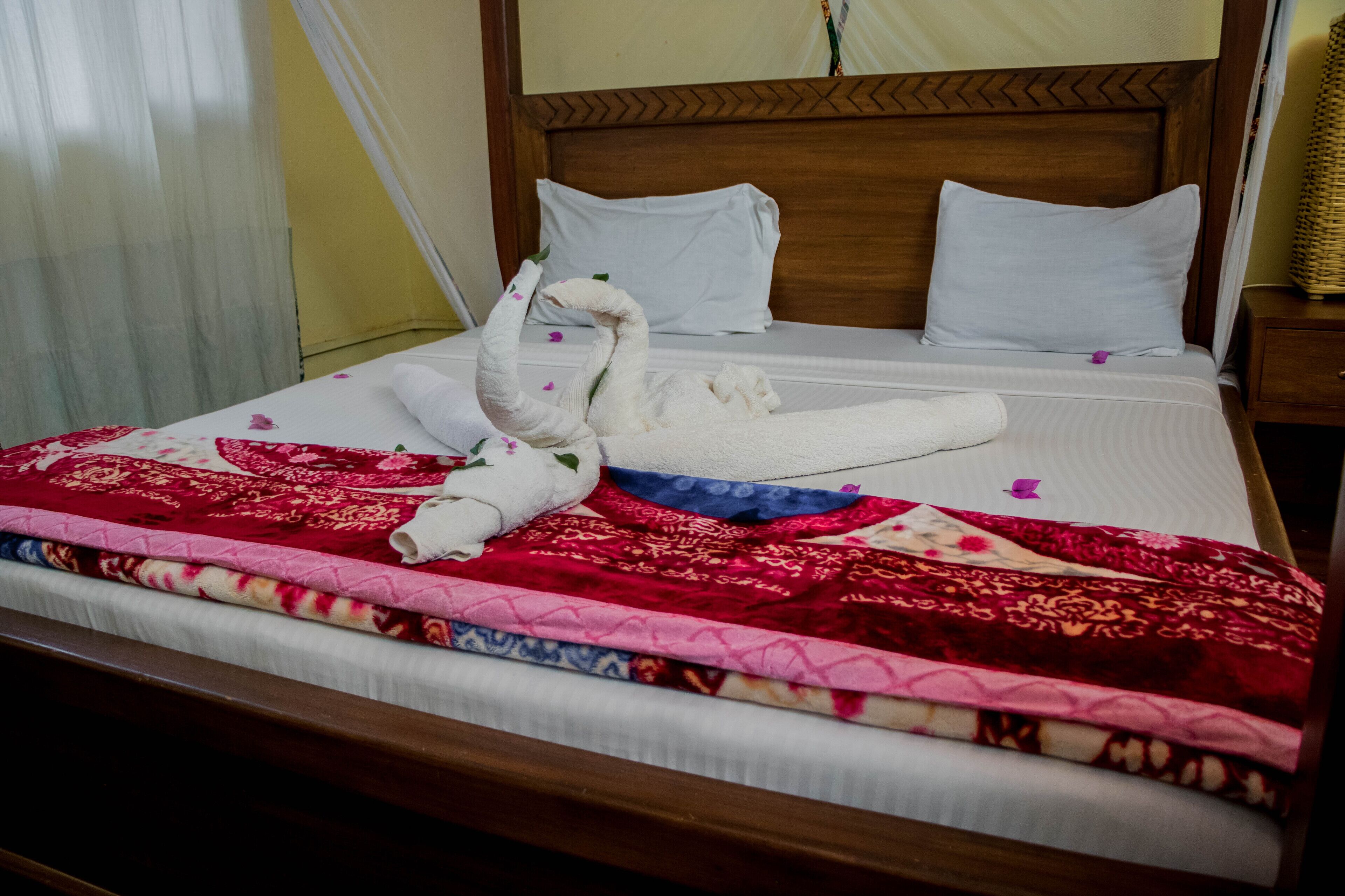 Una cama bien hecha con un arreglo de toallas decorativo con forma de cisne, una colcha estampada en rojo y rosa, y un cabecero de madera con diseño en chevron.