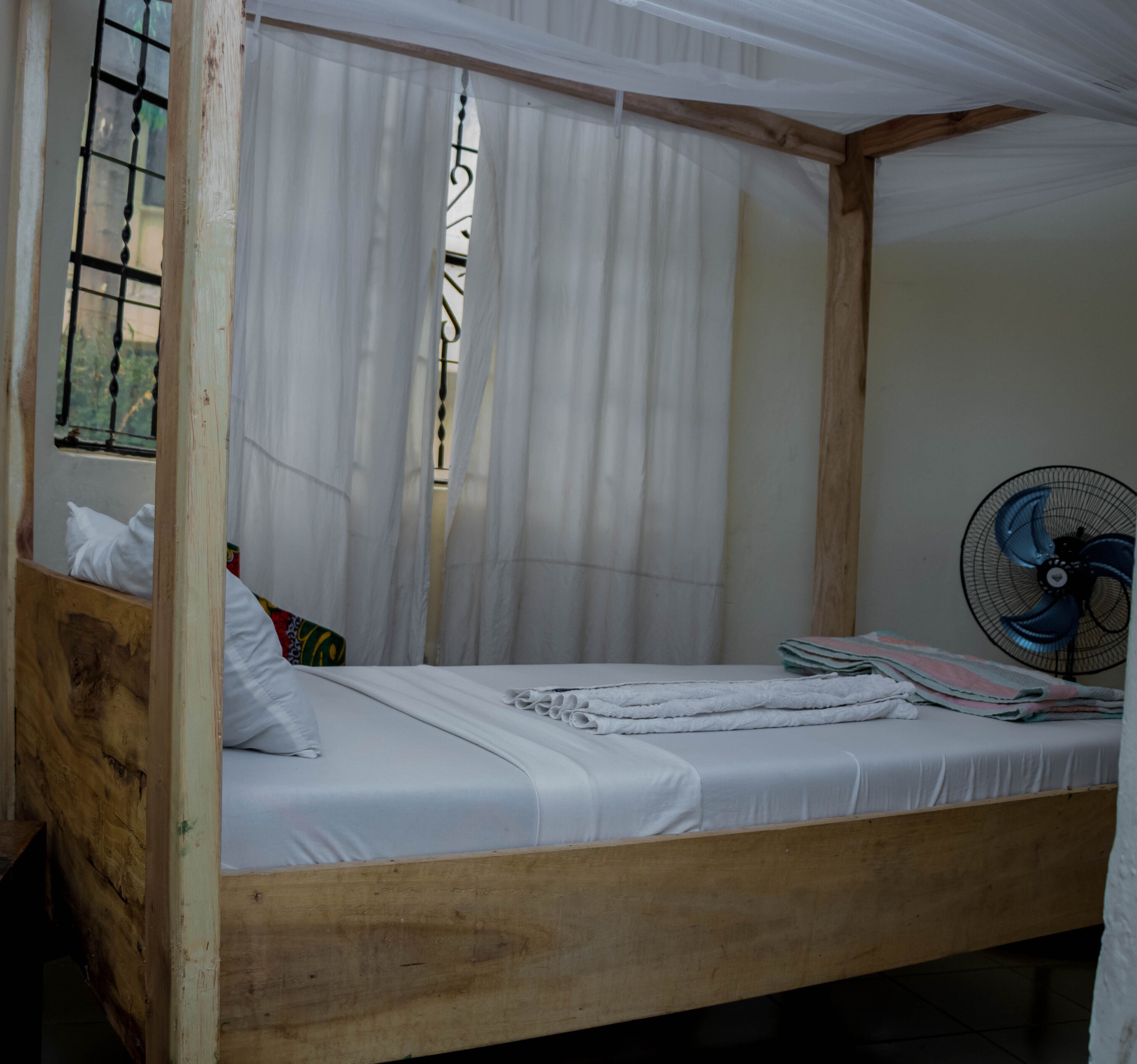 A single-occupancy room with a wooden bed frame, white bedding, a window with metal bars, and a wall-mounted fan.