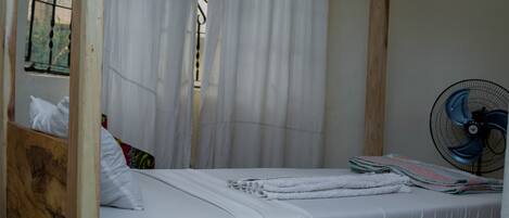 A single-occupancy room with a wooden bed frame, white bedding, a window with metal bars, and a wall-mounted fan.
