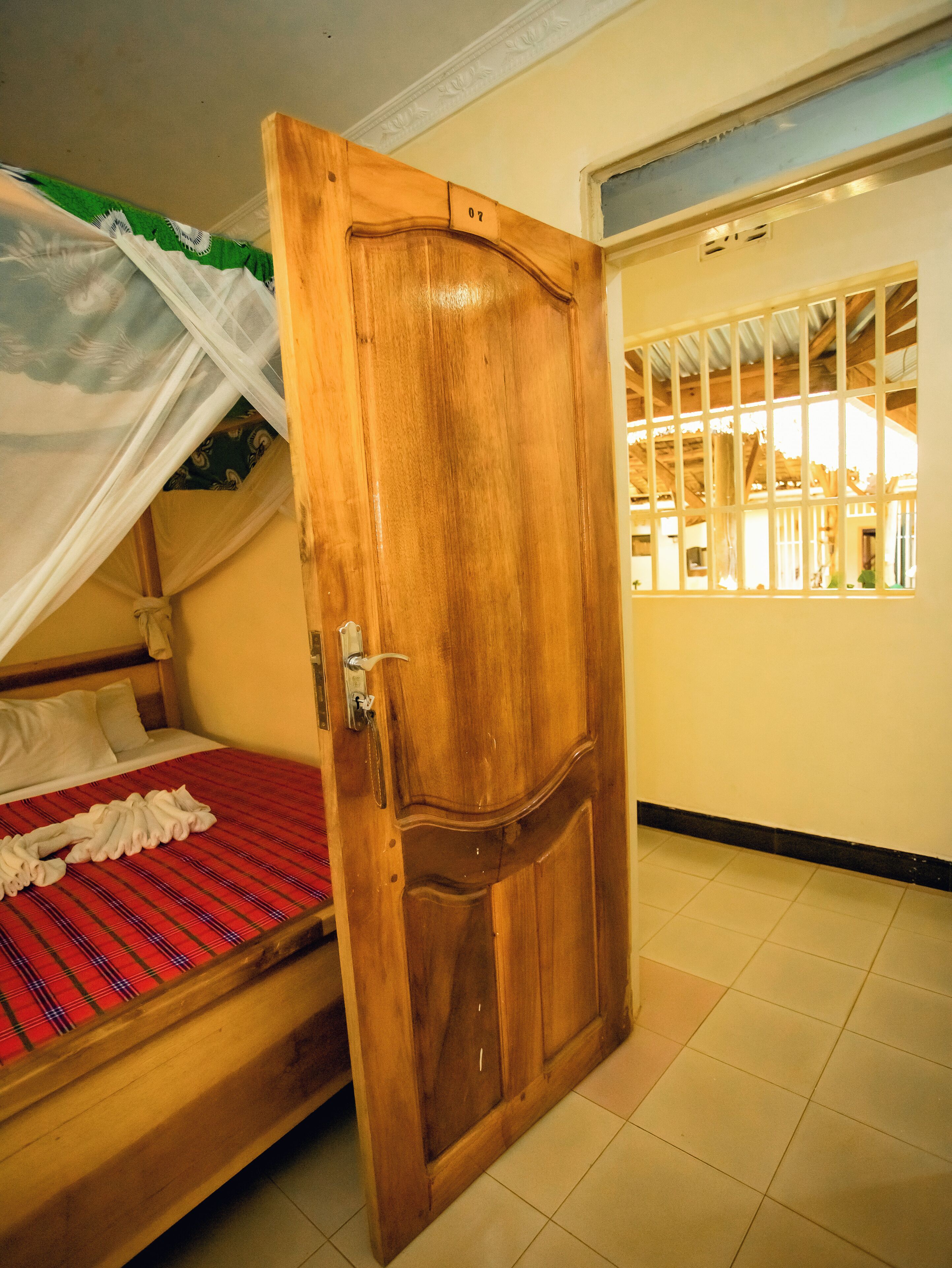 Una porta in legno con un numero, un letto con copripiumino rosso a quadri e una finestra con sbarre.
