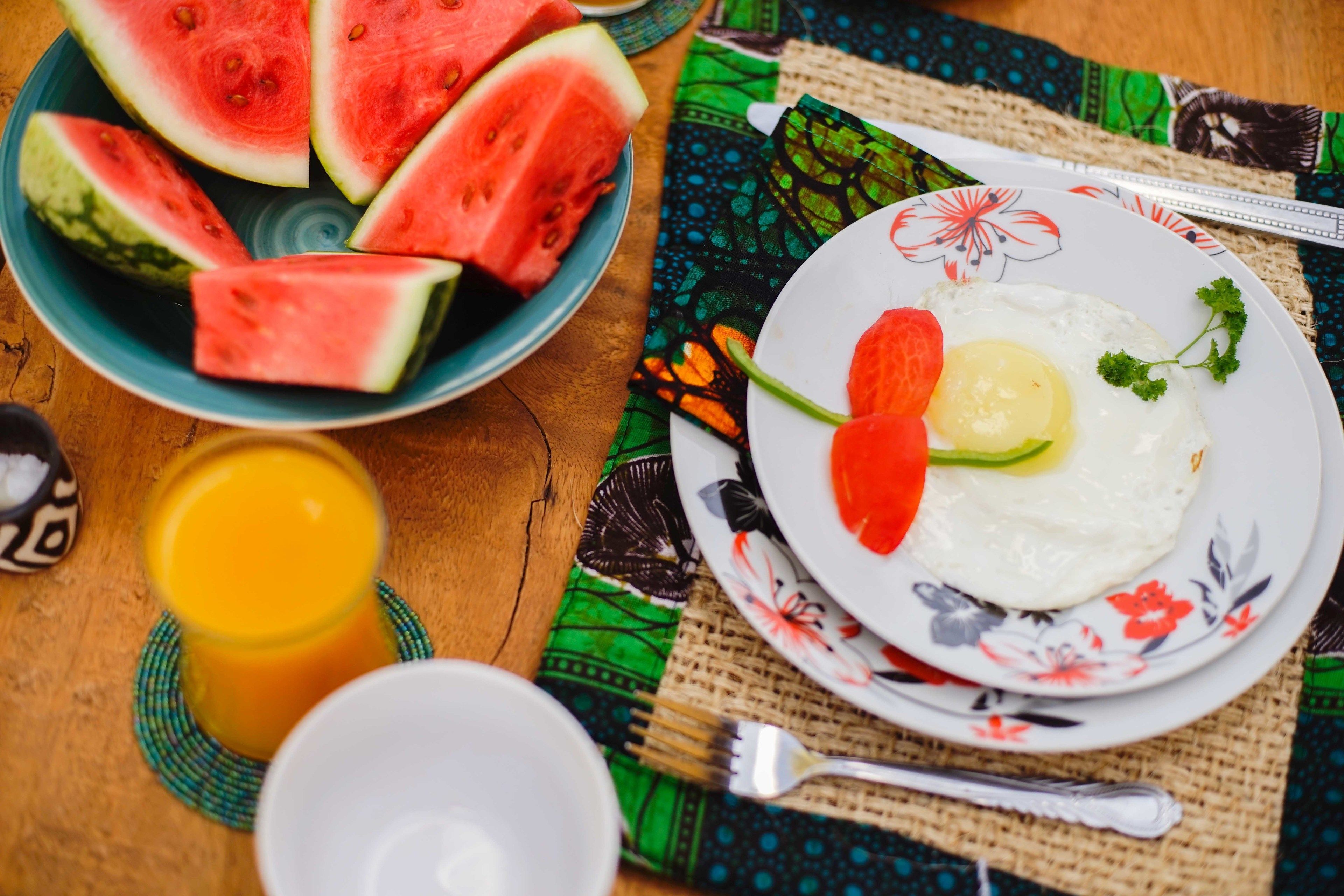 A plate with a fried egg, tomato, and cucumber on a floral plate, a bowl of watermelon slices, and a glass of orange juice on a wooden table with a colorful tablecloth.
