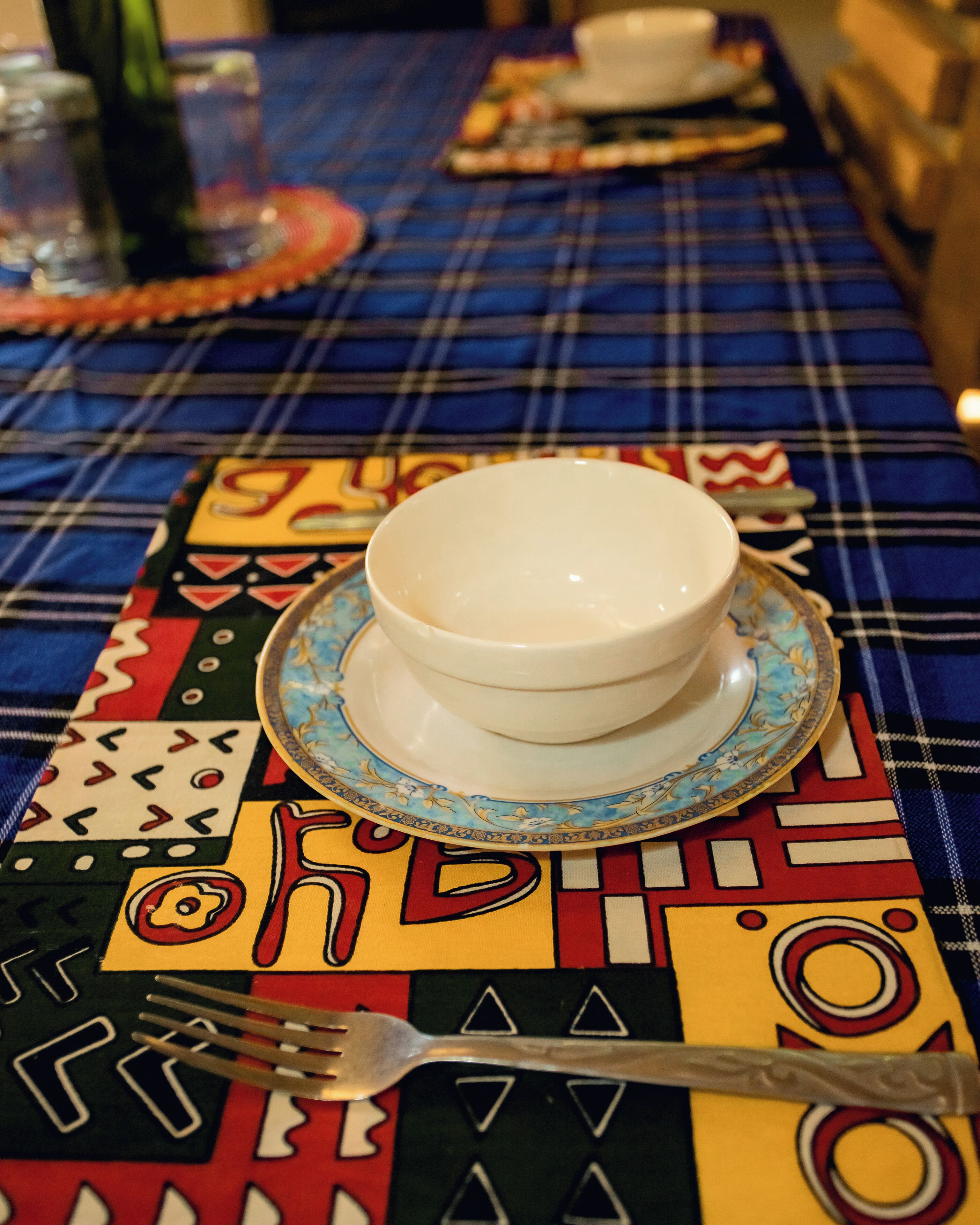 A table with a white bowl on a blue and gold patterned plate, a fork, and a patterned placemat.