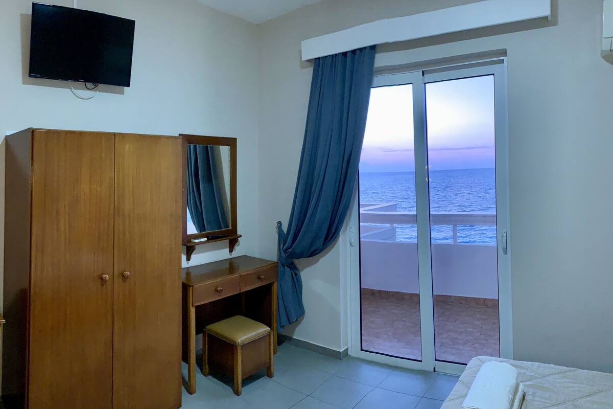 A hotel room with a wooden wardrobe, a small desk with a mirror, a window with a view of the sea, and a bed with a white towel.