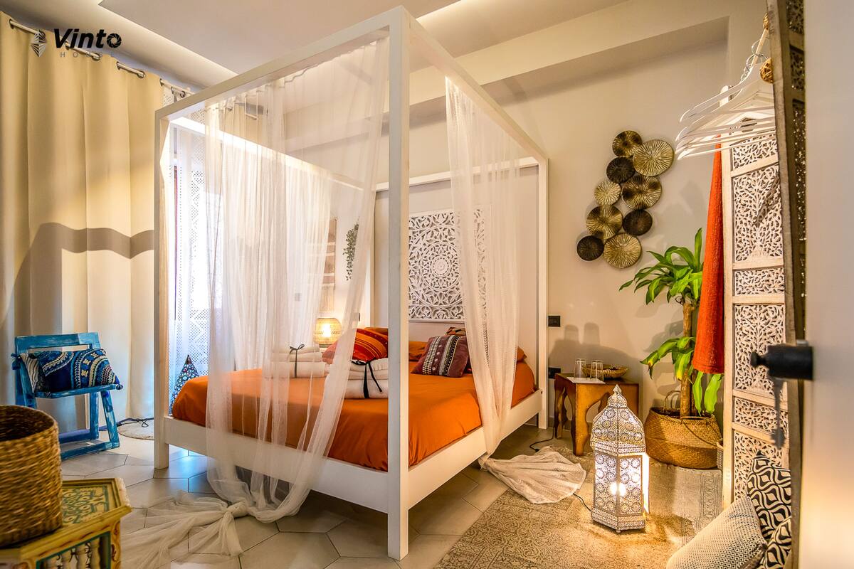 A bedroom with a four-poster bed, a small table with a lamp, a chair, and decorative wall art.